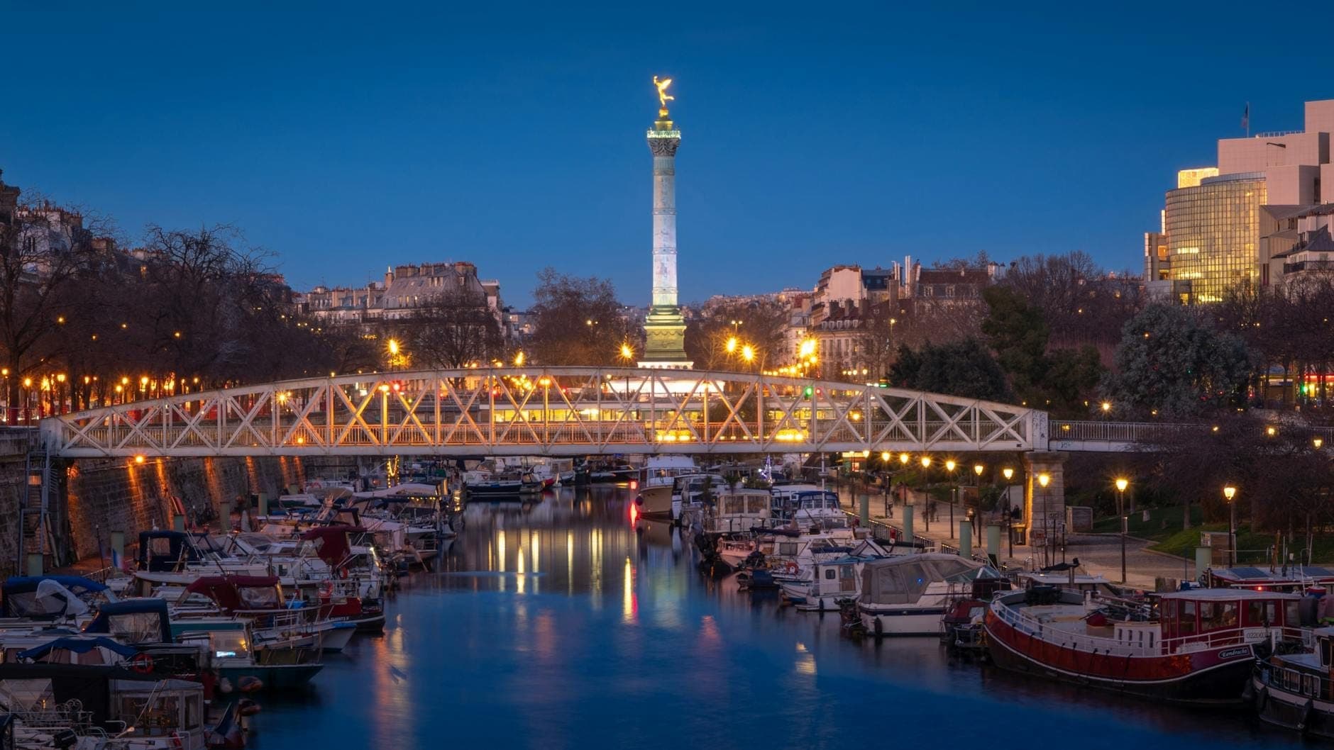 Evening view of the Bastille column and marina with boats docked along the water, a pedestrian bridge in the foreground, and city lights reflecting on the canal.
