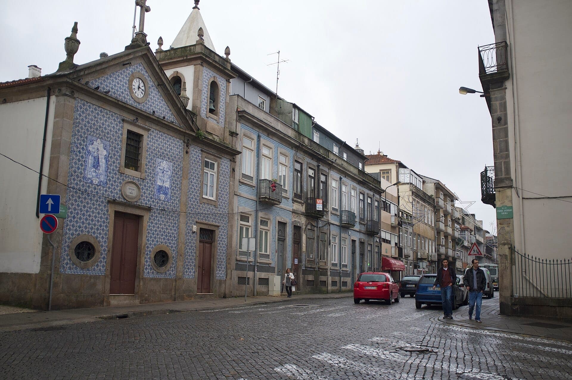 Eine mit Azulejos bedeckte Kirche und traditionelle Gebäude säumen eine ruhige, nasse Straße in Batalha, Porto, mit einigen Fußgängern und geparkten Autos.