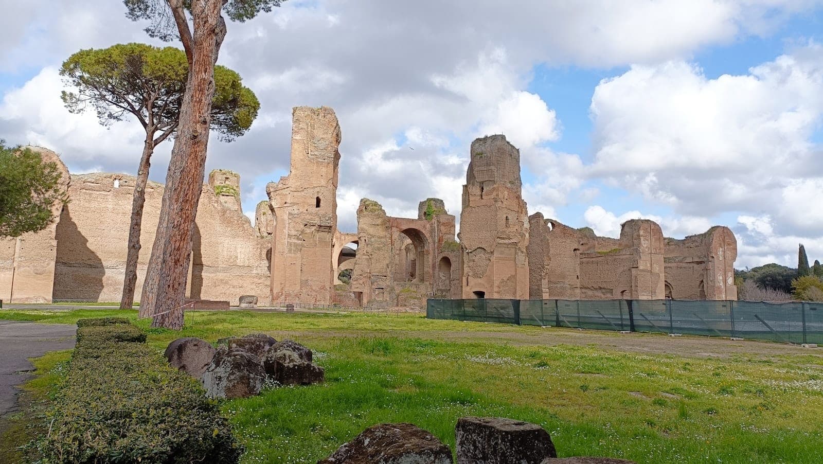 Le rovine delle Terme di Caracalla a Roma, con imponenti mura di mattoni antichi, pietre sparse, alberi e un cielo suggestivo sullo sfondo.