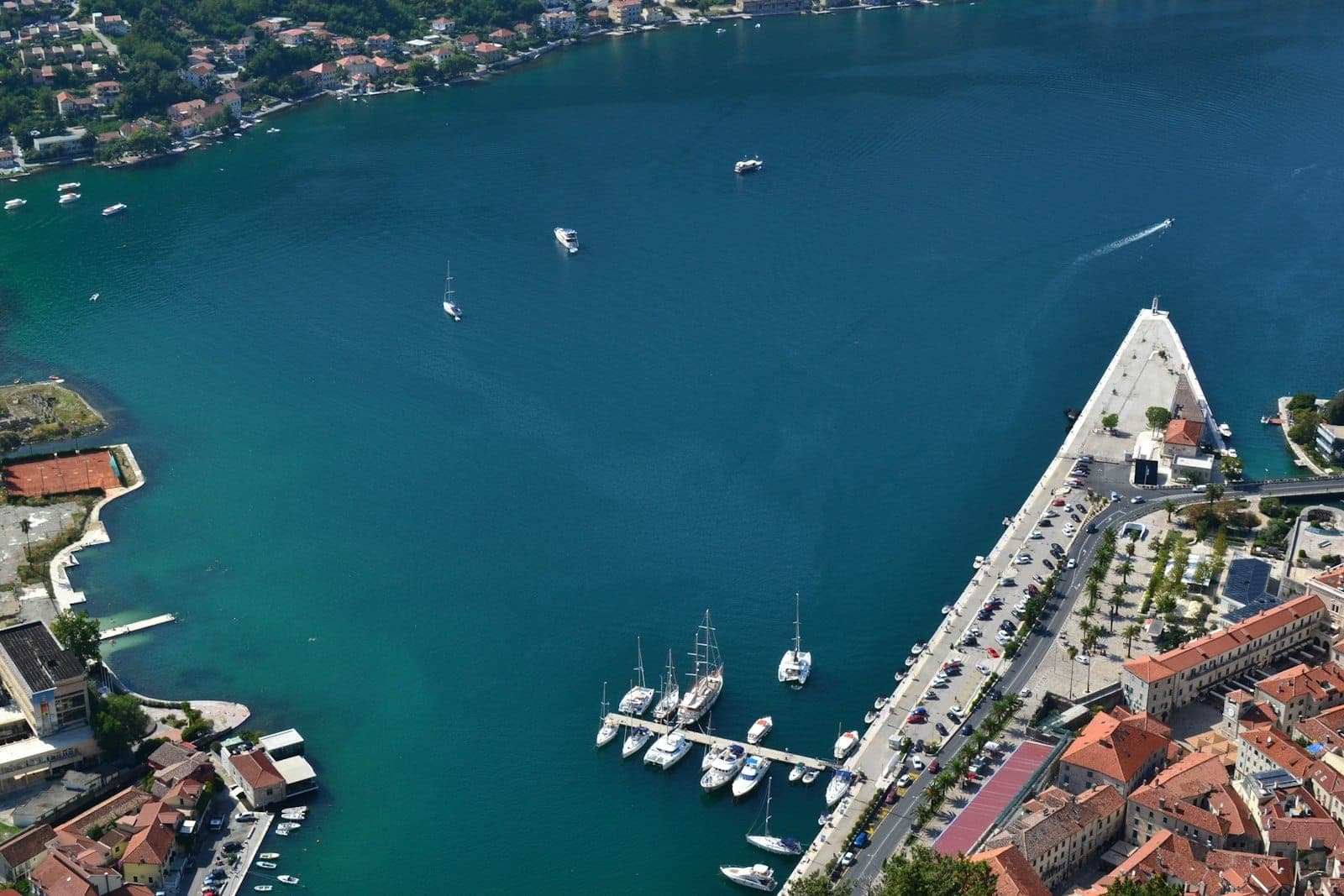 Vue aérienne de la marina de Kotor avec plusieurs bateaux amarrés, adjacente aux remparts de la vieille ville et à la promenade en bord de mer.