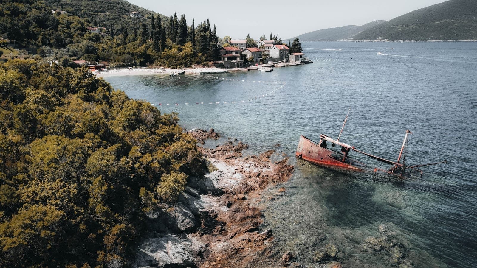 Vue aérienne d'une petite baie avec un bateau naufragé près d'un rivage rocheux et un village en arrière-plan le long du littoral de la Baie de Kotor.