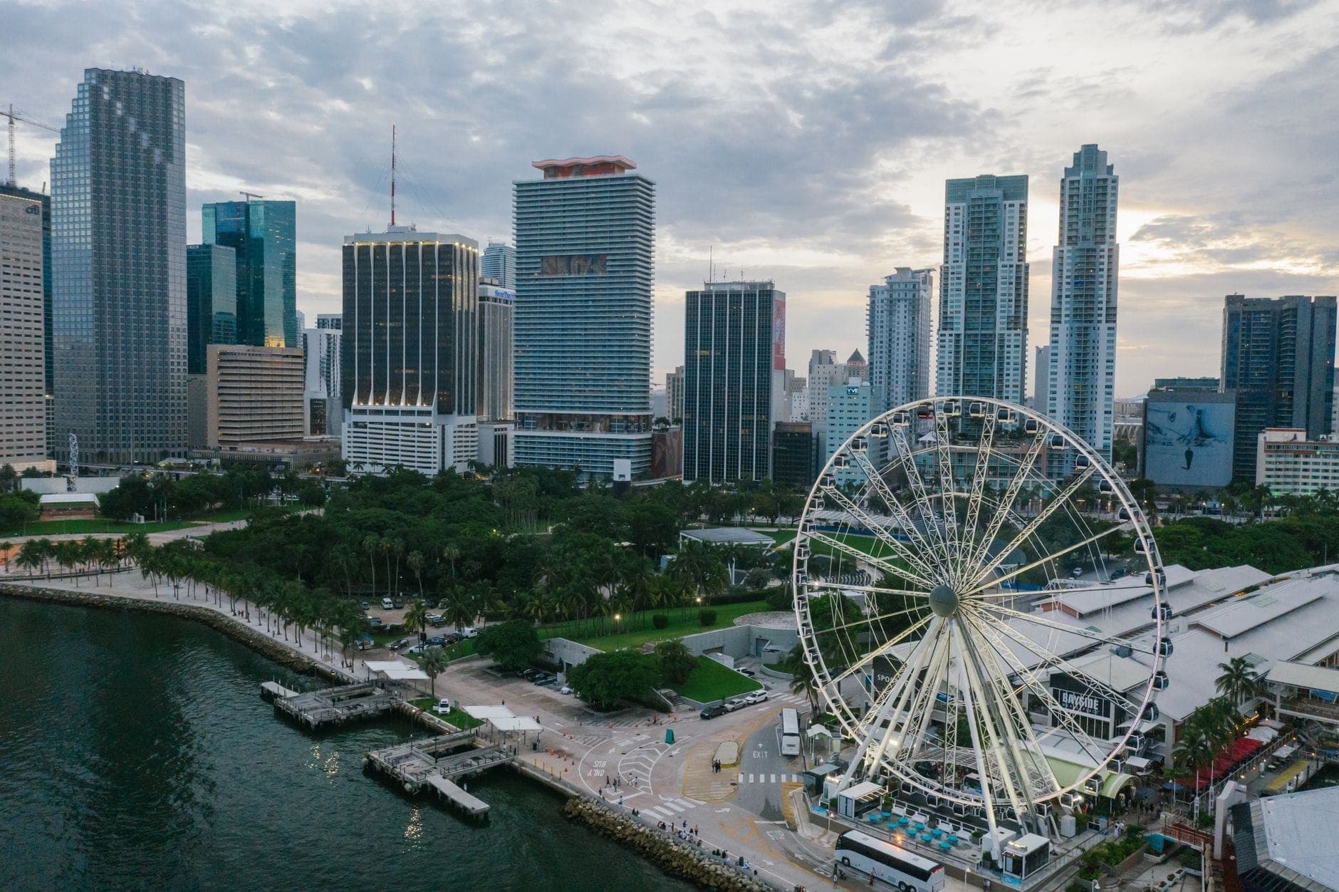 Aerial view of Bayfront Park with palm trees, waterfront paths, Ferris wheel, and Miami skyscrapers in the background on a partly cloudy day.
