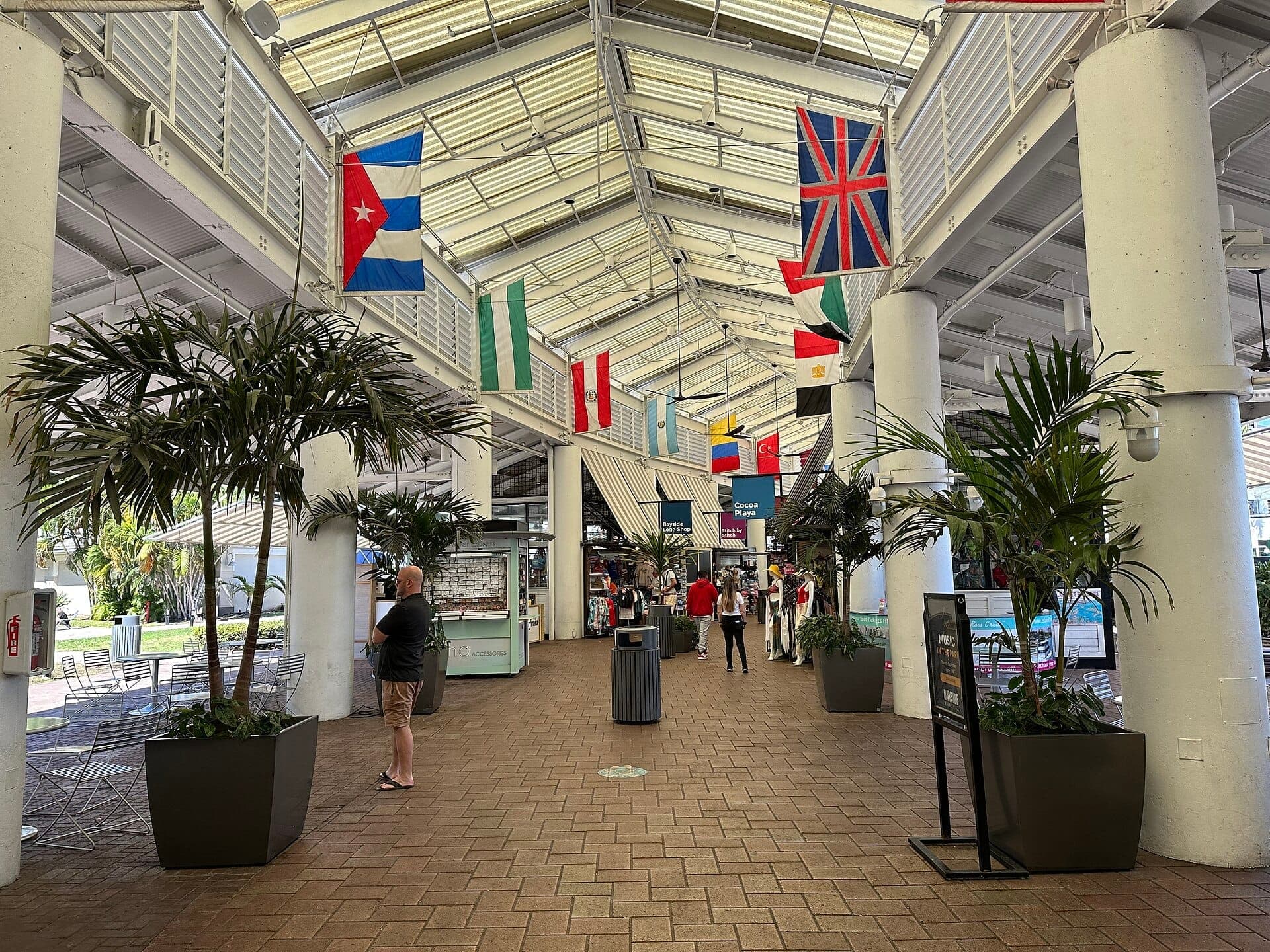 Couloir intérieur du Bayside Marketplace avec des drapeaux internationaux, des plantes tropicales et plusieurs personnes faisant leurs achats sous un toit en verre lumineux et ensoleillé.