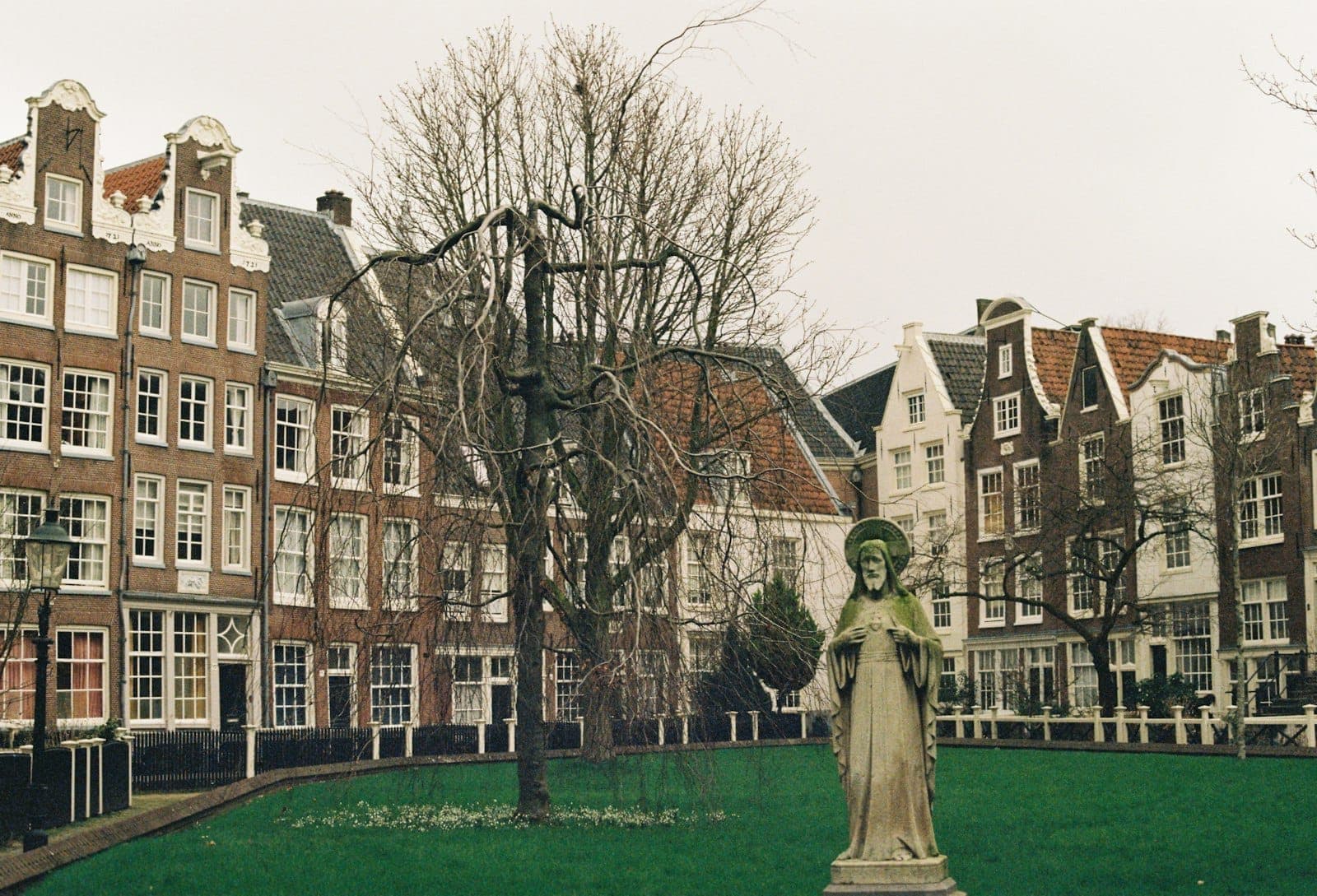 Reihe historischer niederländischer Häuser und eine Statue im zentralen Garten des Begijnhof Amsterdam, mit kahlen Bäumen unter einem bedeckten Himmel.
