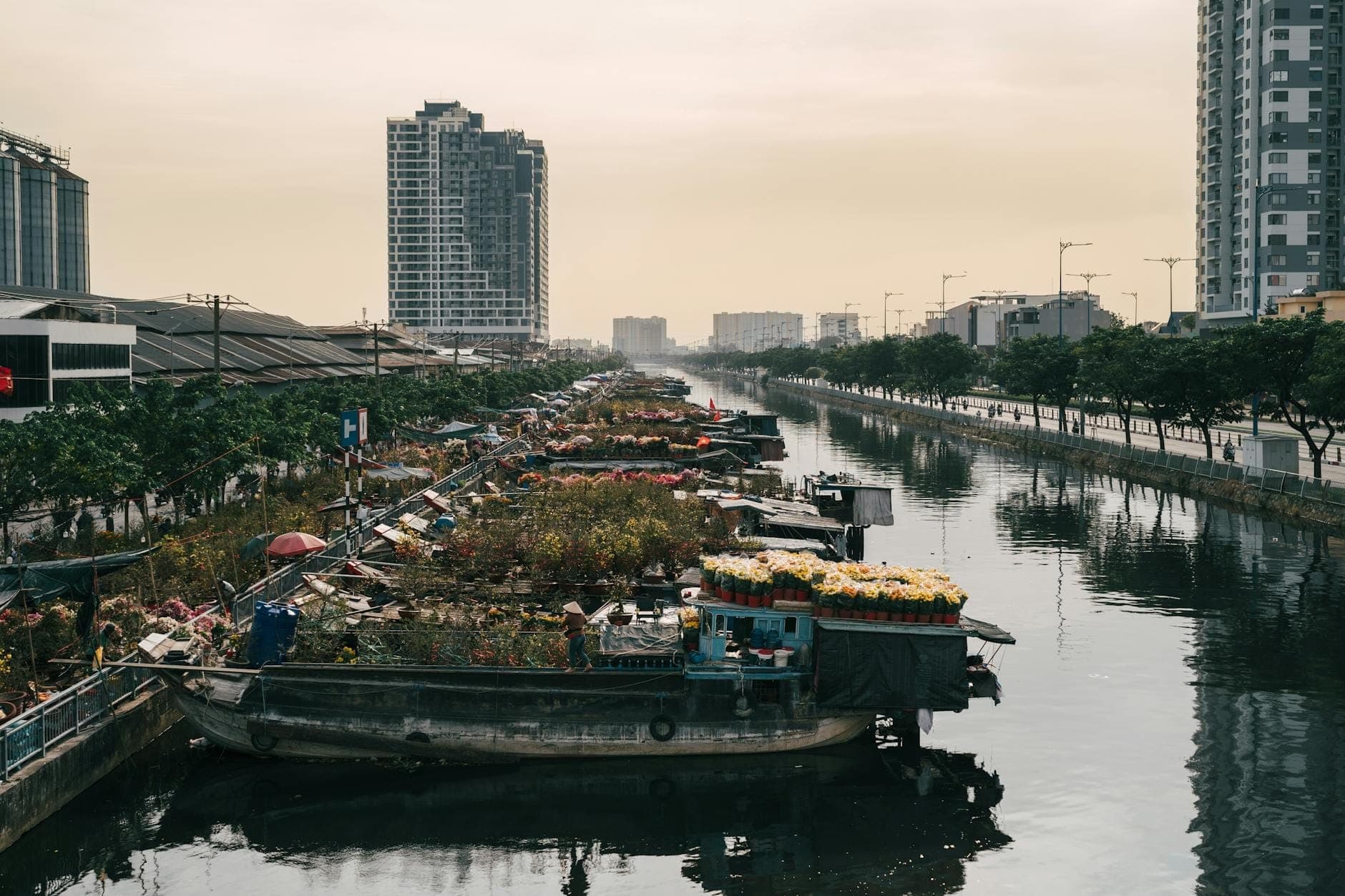 Isang tahimik na kanal sa lungsod na may mga bangkang nagbebenta ng bulaklak at iba pang mga kalakal, napapalibutan ng mga modernong gusali at mga landas na may puno sa dapit-hapon sa Ho Chi Minh City.