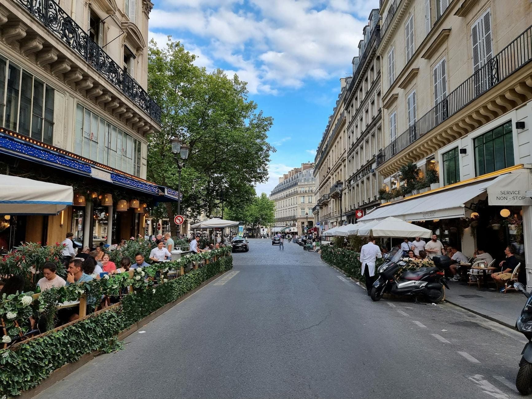Paris street scene with lively outdoor cafes, people dining and relaxing on terraces, tree-lined buildings creating a vibrant, inviting pedestrian atmosphere.