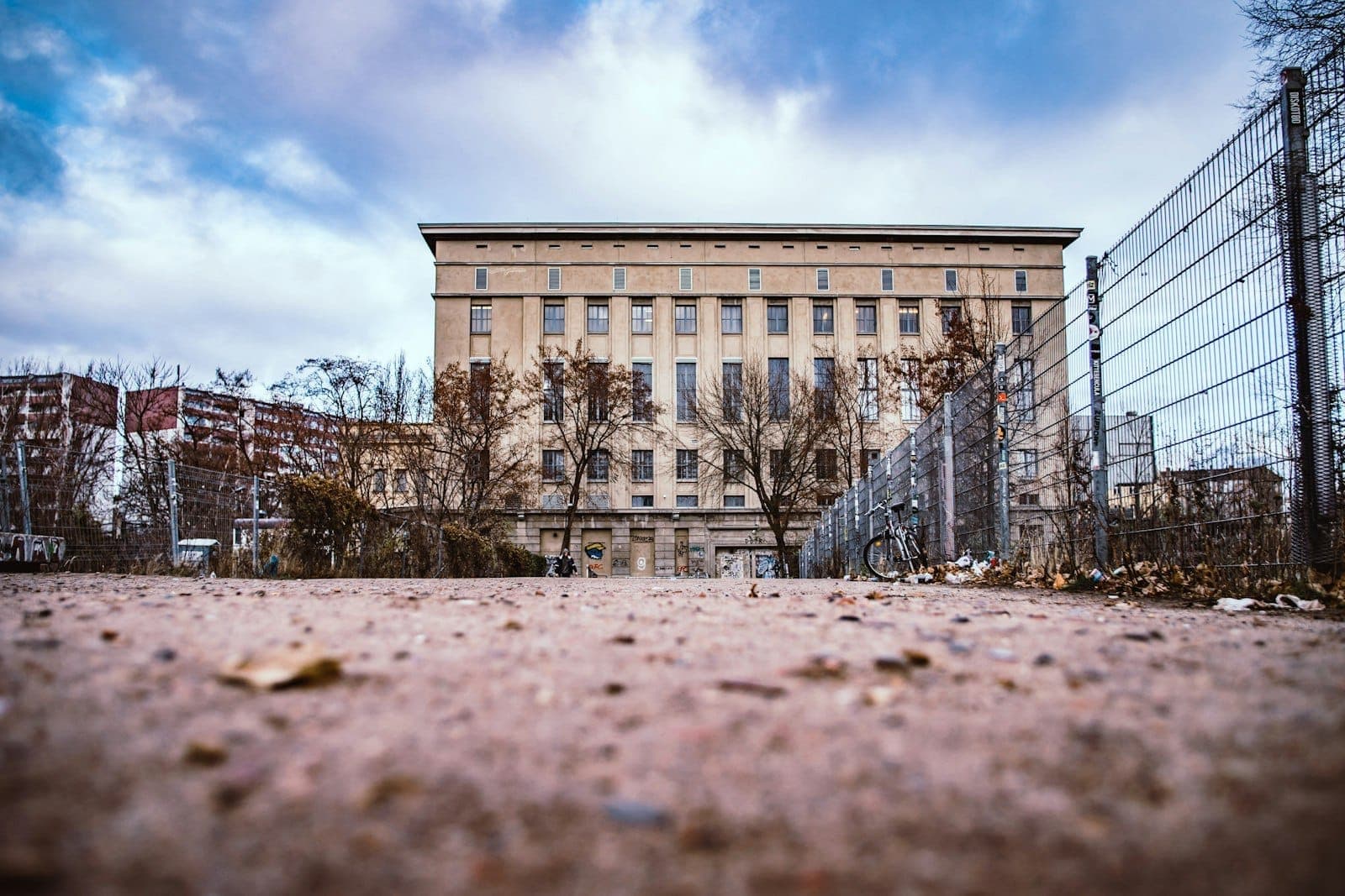 มุมมองกว้างของด้านหน้าอาคารอุตสาหกรรมของ Berghain ท่ามกลางท้องฟ้าครึ้ม มีทางเดินที่มีรั้วกั้น ต้นไม้ไม่มีใบ และจักรยานที่ทางเข้า
