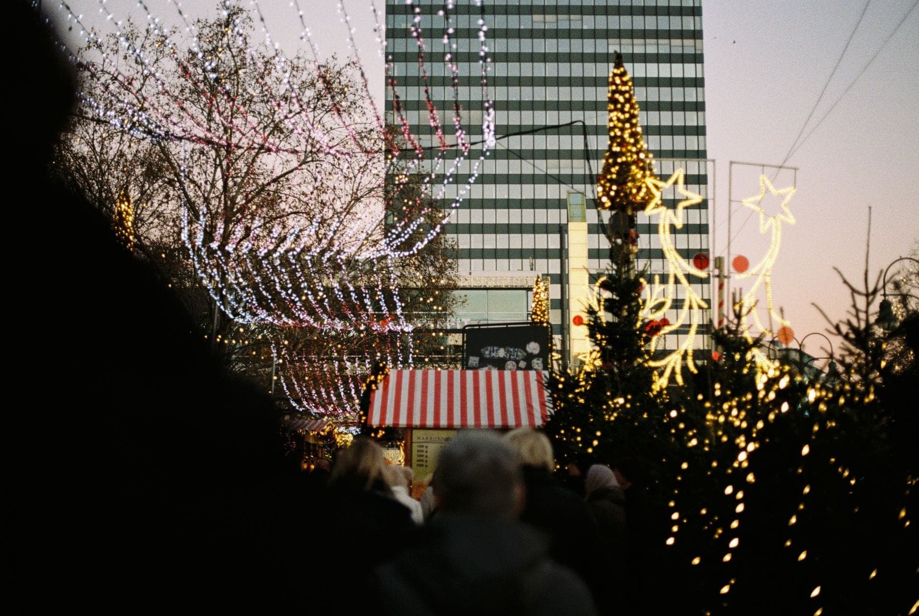 Menschen schlendern durch einen festlichen Berliner Weihnachtsmarkt mit dekorierten Ständen, funkelnden Lichtern und einem modernen Hochhaus im Hintergrund.