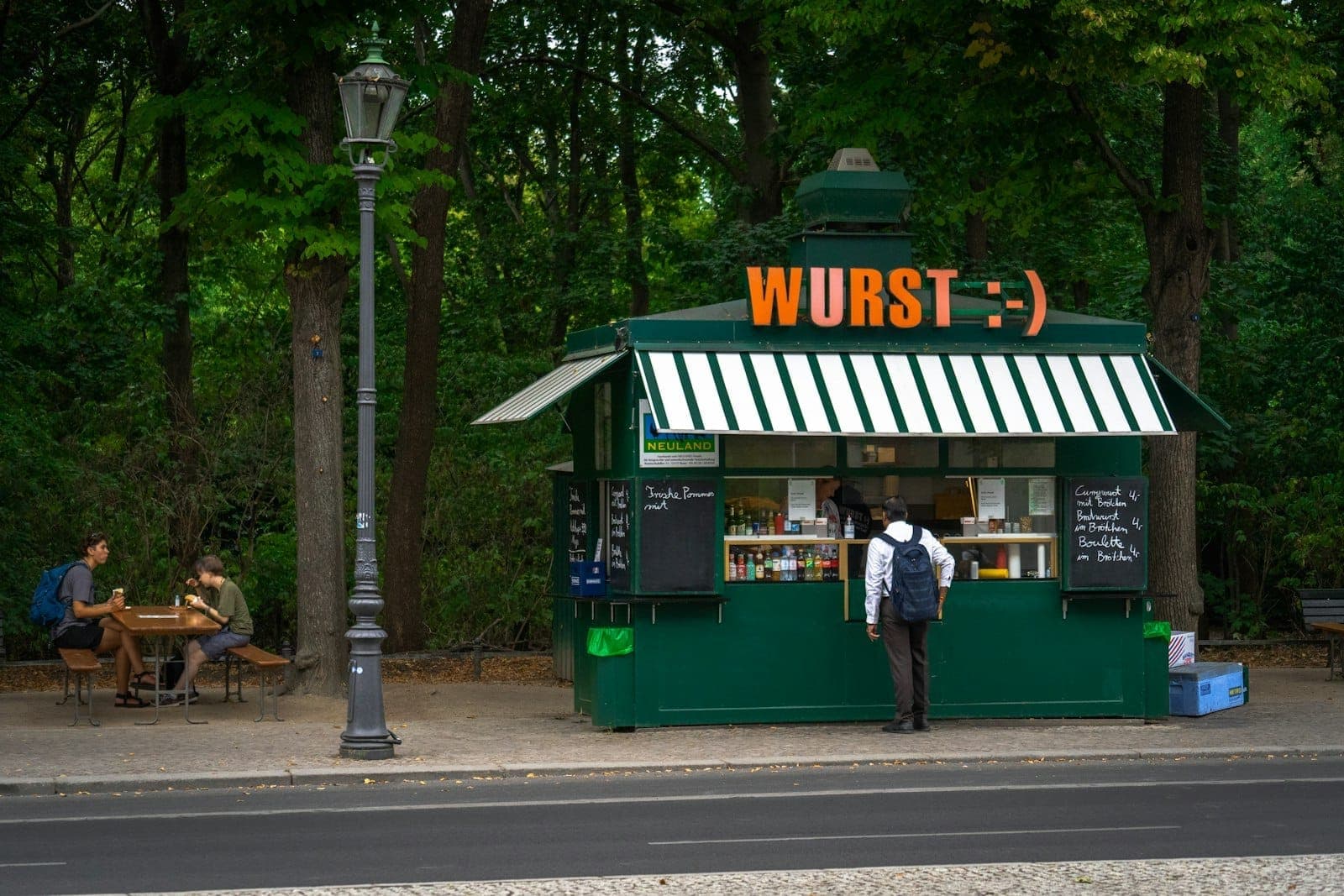 ภาพซุ้มอาหารข้างถนนสีเขียวในเบอร์ลินชื่อ WURST มีคนต่อแถวสั่งอาหารและนั่งกินที่โต๊ะกลางแจ้งใต้ร่มไม้