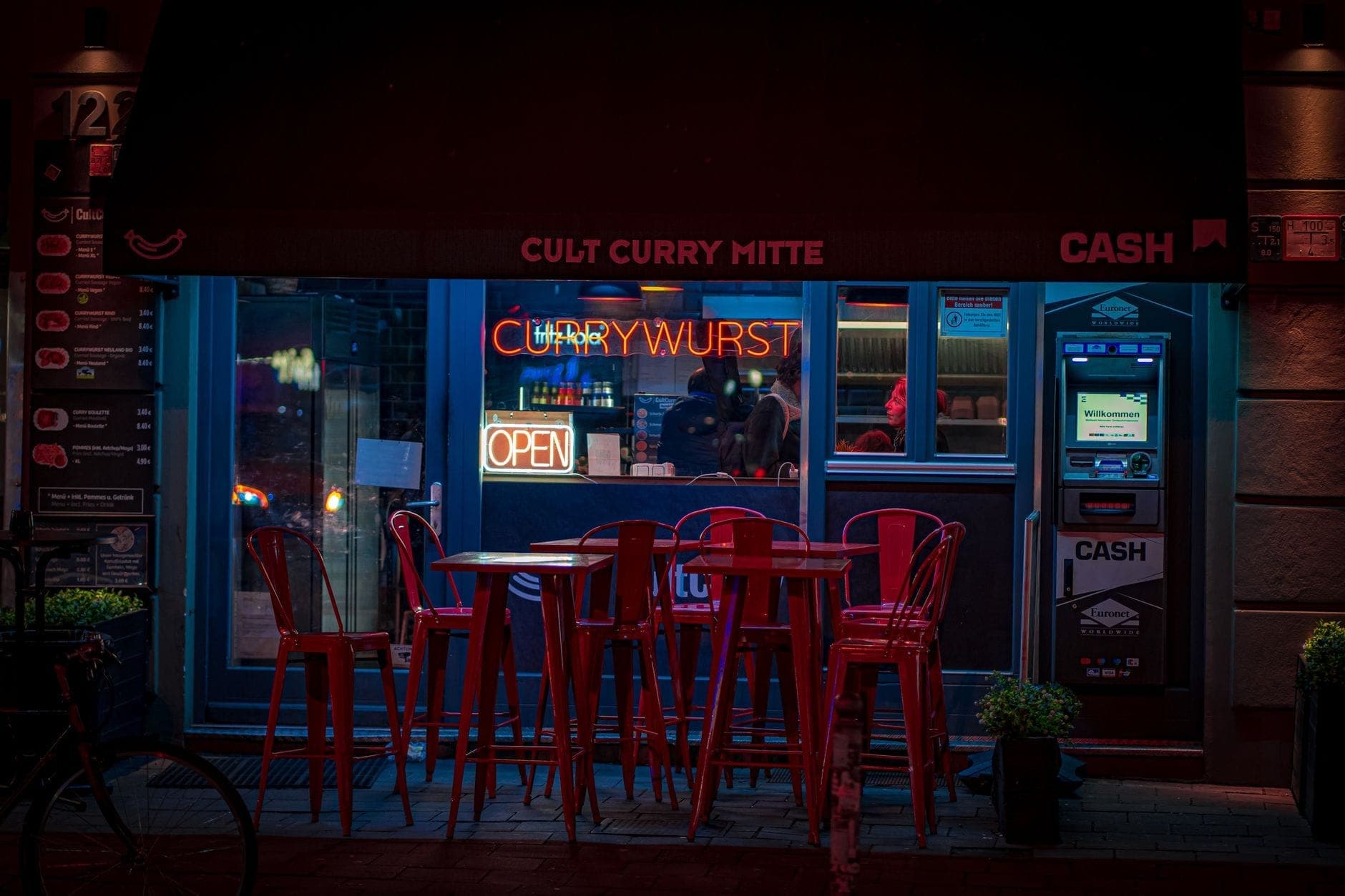 Ночной вид на уличный фуд-стенд в Берлине с неоновой вывеской 'CURRYWURST', красными стульями и столами и людьми внутри.