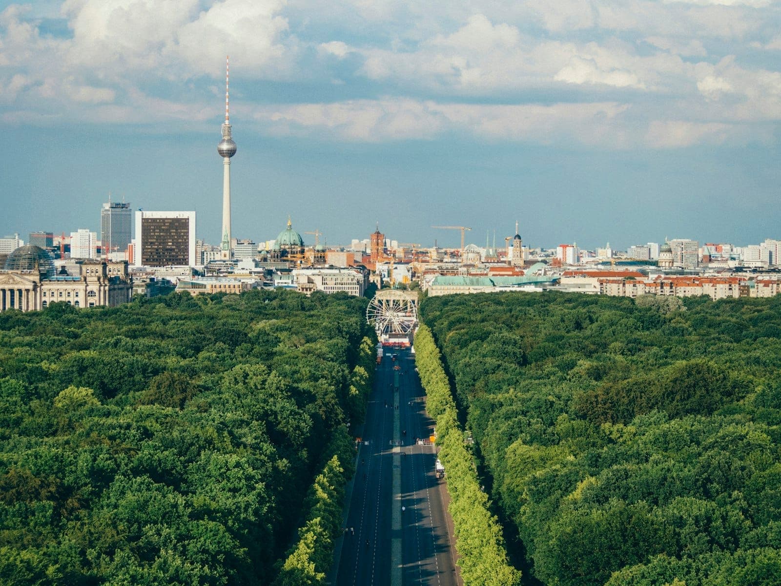Vista aérea sobre el parque Tiergarten de Berlín con densos árboles verdes, el horizonte de la ciudad al fondo y la Torre de Televisión visible, bajo un cielo parcialmente nublado.