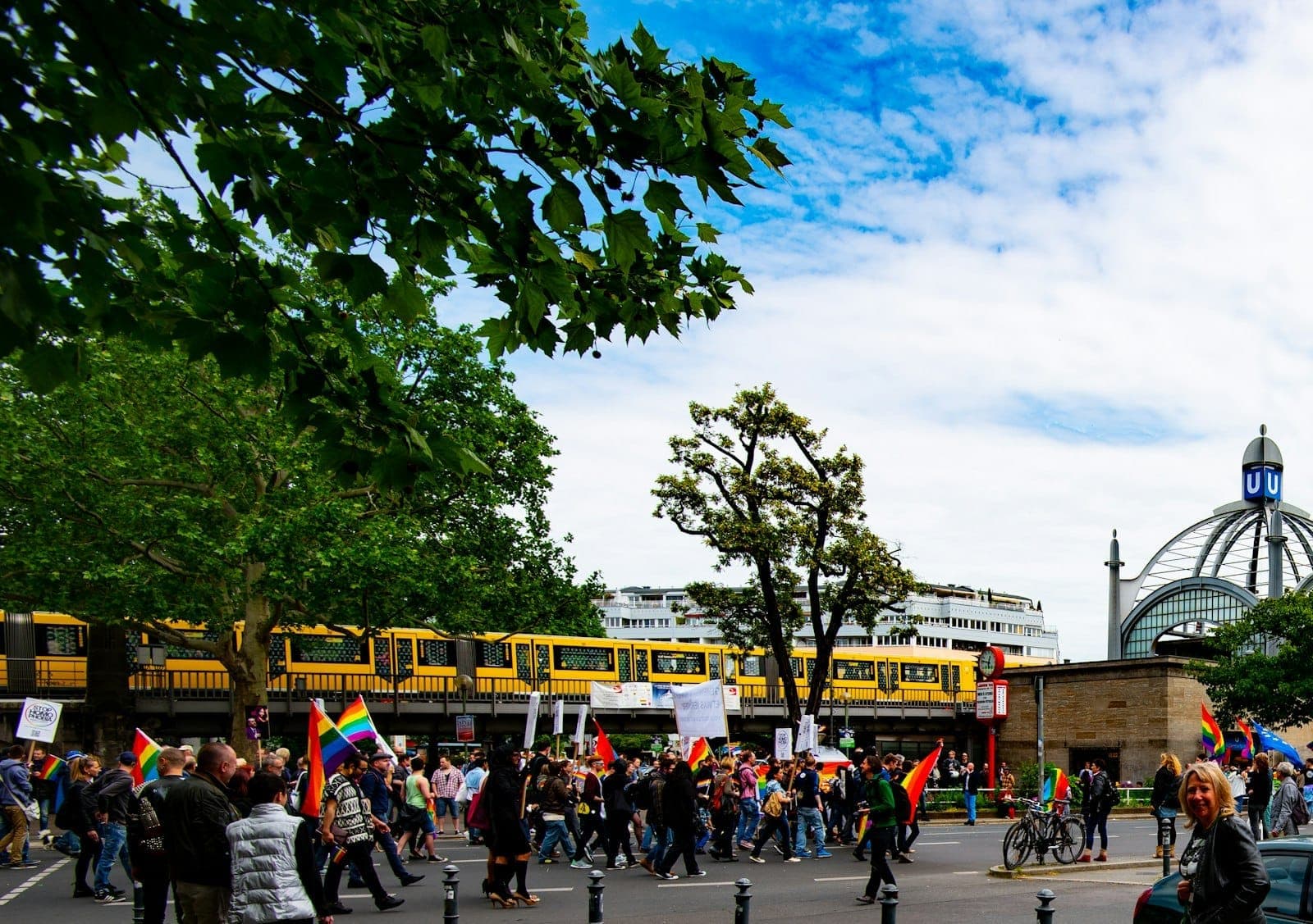 Un animado desfile callejero en Berlín con banderas arcoíris, multitudes y un reconocible tren amarillo de la U-Bahn de Berlín en lo alto, árboles y arquitectura urbana.