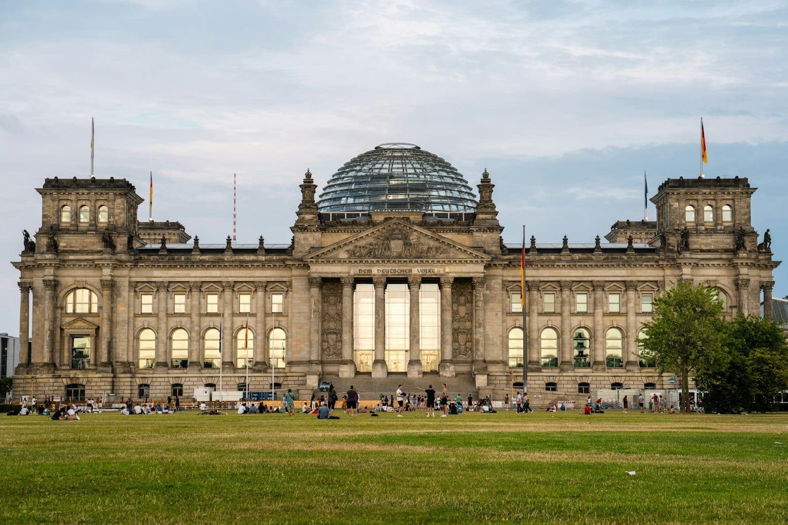 Vista amplia del edificio Reichstag de Berlín con su cúpula de cristal y grandes multitudes de verano en el césped frente a él bajo un cielo despejado.