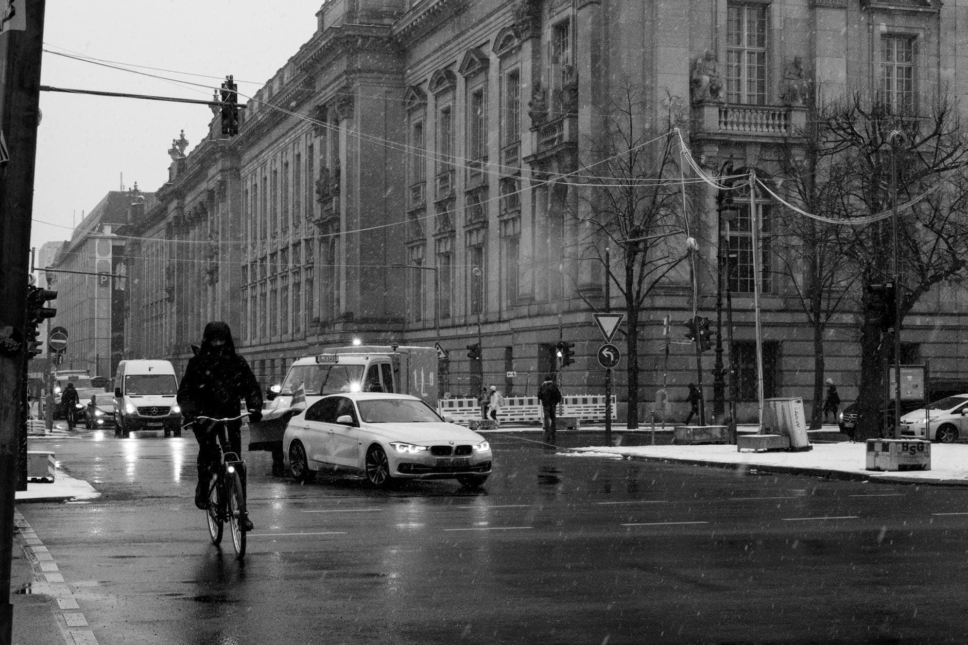 Berliner Straße im Winter mit leichtem Schnee auf dem Boden, Radfahrer, Autos und prächtigen historischen Gebäuden unter bedecktem Himmel.