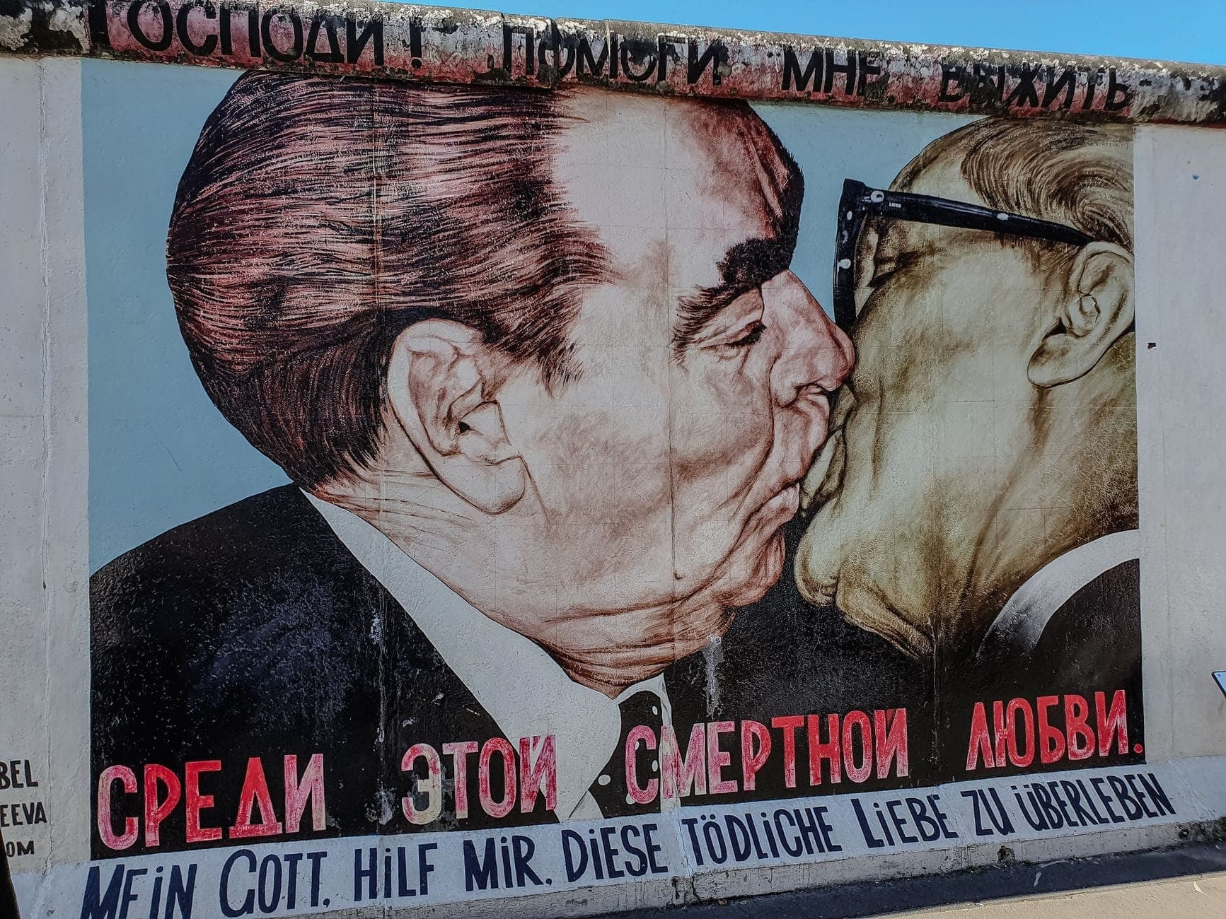 Close-up of the famous mural of two men kissing on a preserved section of the Berlin Wall under blue sky.