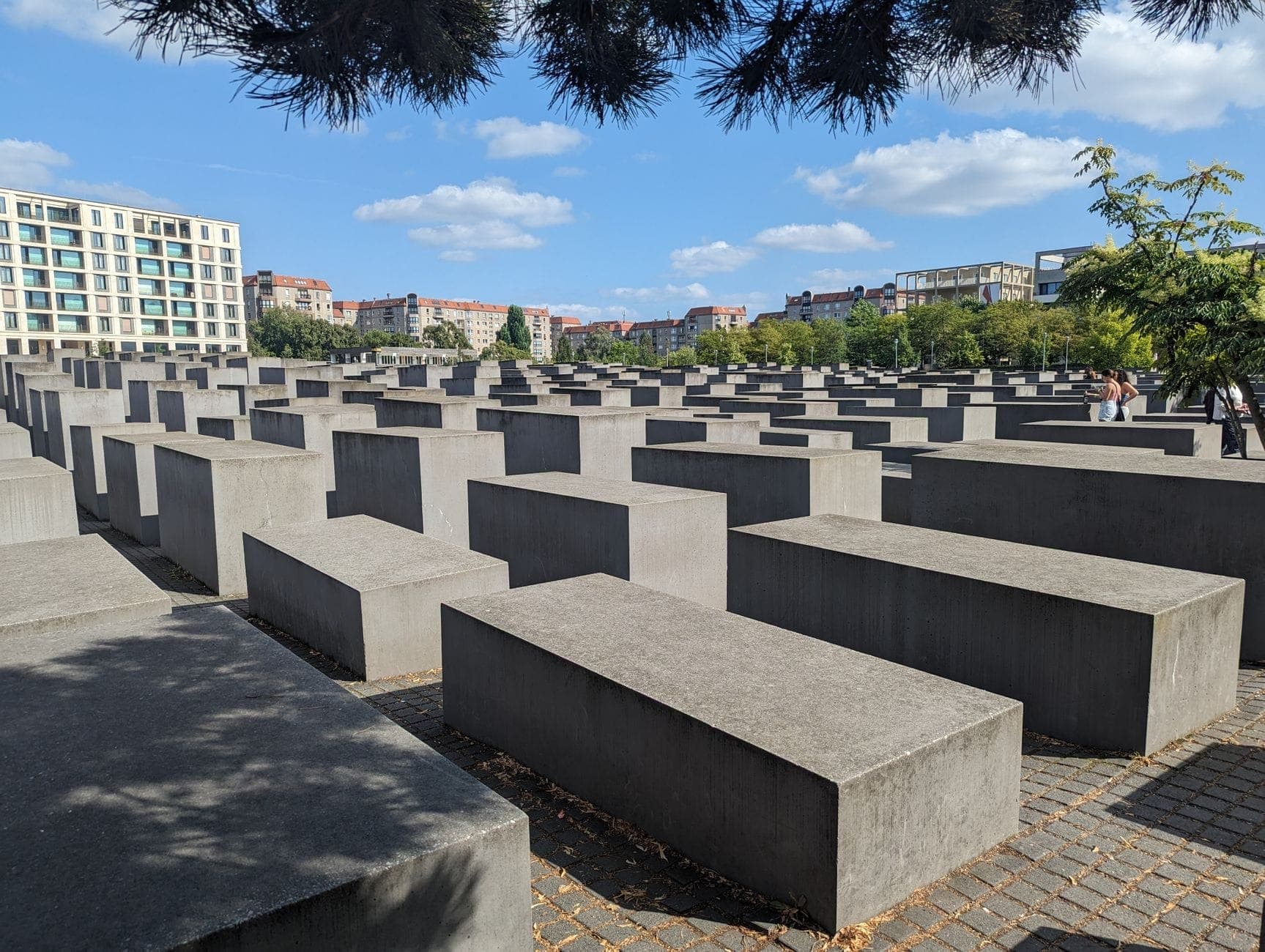 Photo of Berlin's Holocaust Memorial, showing rows of concrete slabs with city buildings and blue sky in the background.