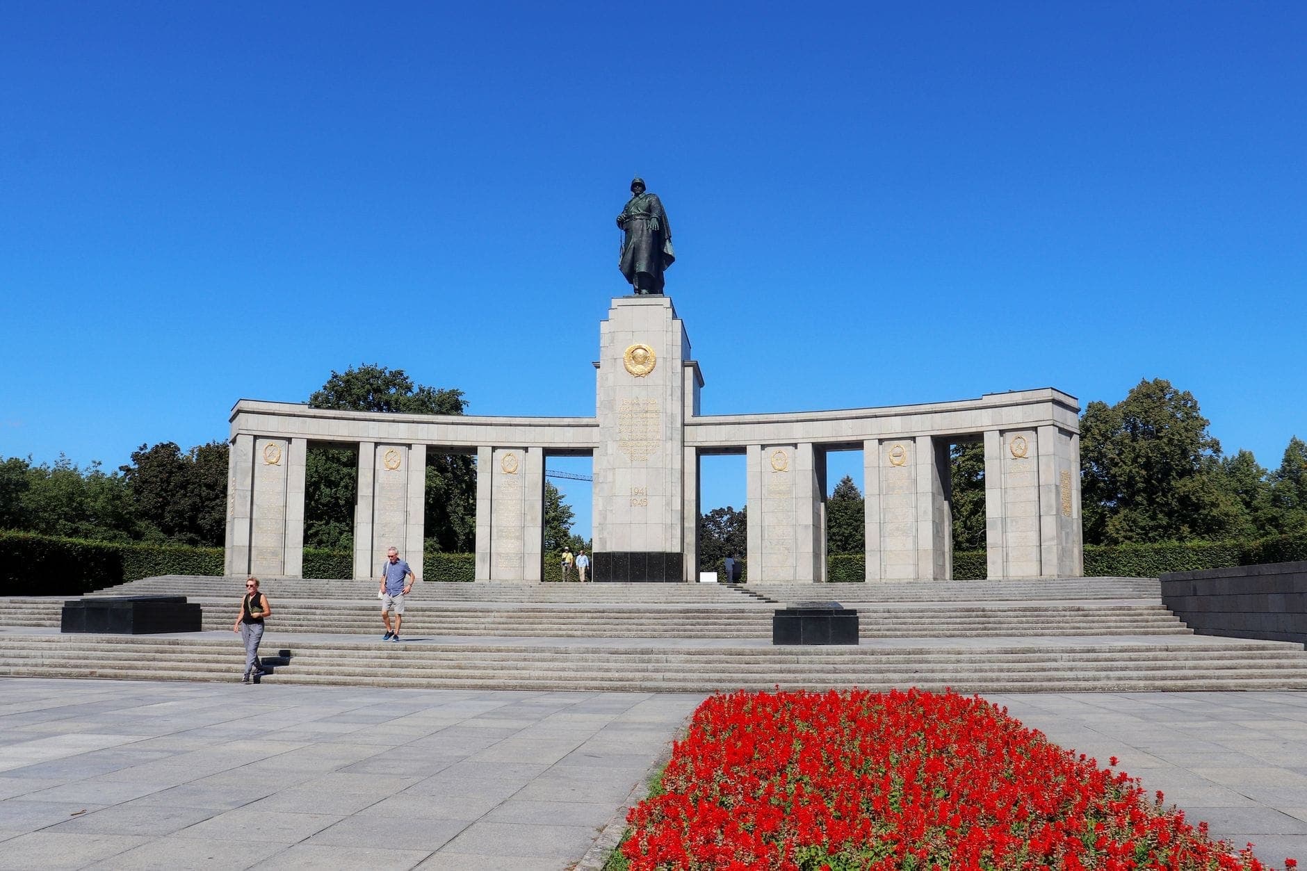 Wide view of Berlin's Soviet War Memorial in Tiergarten, with a statue, columns, steps, and red flowerbeds on a sunny day.