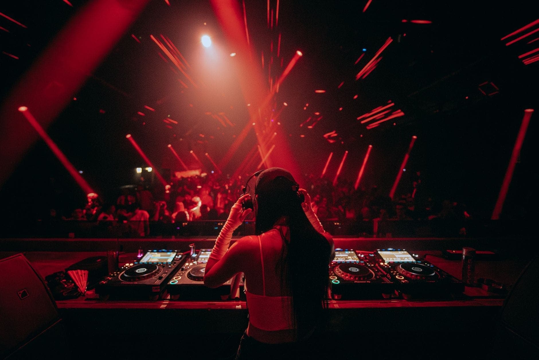 Ein DJ bei einem Auftritt in einem Club mit roter Beleuchtung, Blick von hinter dem DJ-Pult, vollbesetzte Tanzfläche im Hintergrund