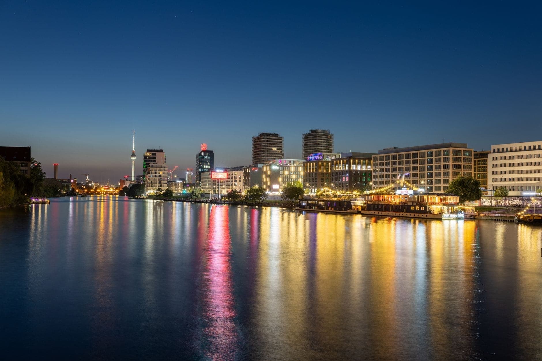 Nachtansicht der Spree in Berlin mit bunten Spiegelungen, beleuchteten Gebäuden und der Skyline mit dem Fernsehturm im Hintergrund.