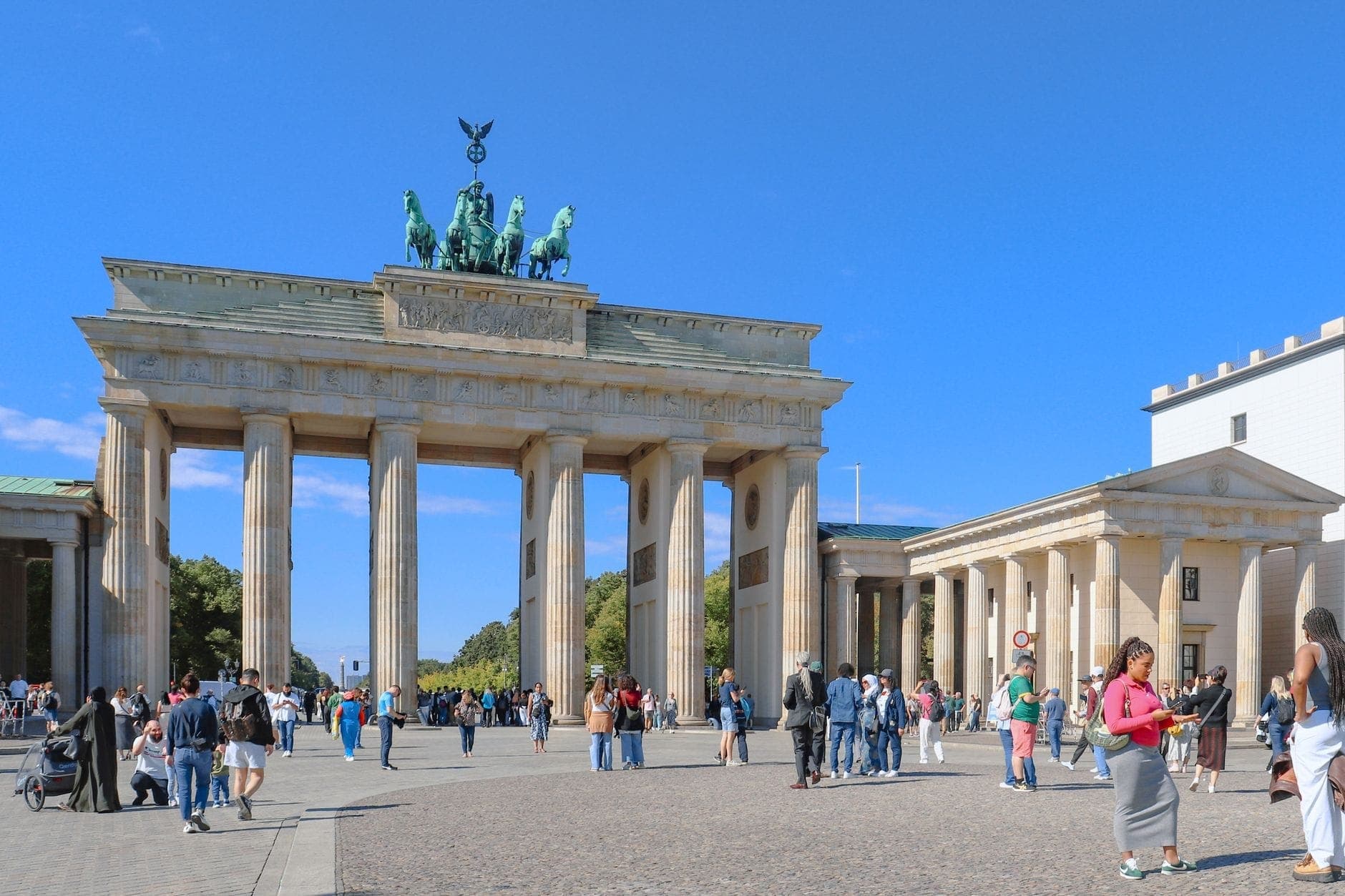 Una piazza affollata a Berlino con persone che camminano e si radunano davanti alla famosa Porta di Brandeburgo sotto un cielo azzurro limpido.