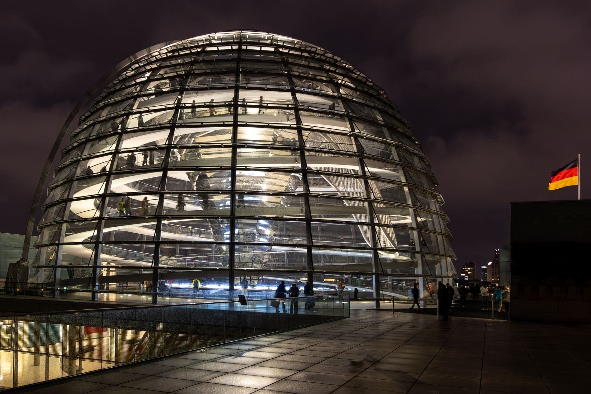 Cupola del Reichstag illuminata di notte con persone visibili all'interno, bandiera tedesca e cielo nuvoloso sullo sfondo.