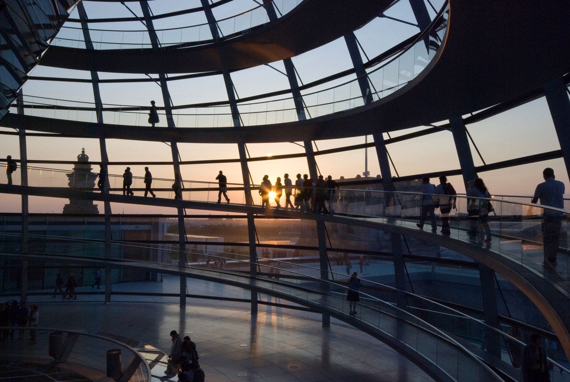 ผู้เข้าชมเดินอยู่ภายในโดมกระจก Reichstag ยามพระอาทิตย์ตก แสงธรรมชาติอ่อน ๆ ส่องทางเดินโค้งและฉากหลังเป็นวิวกรุงเบอร์ลิน