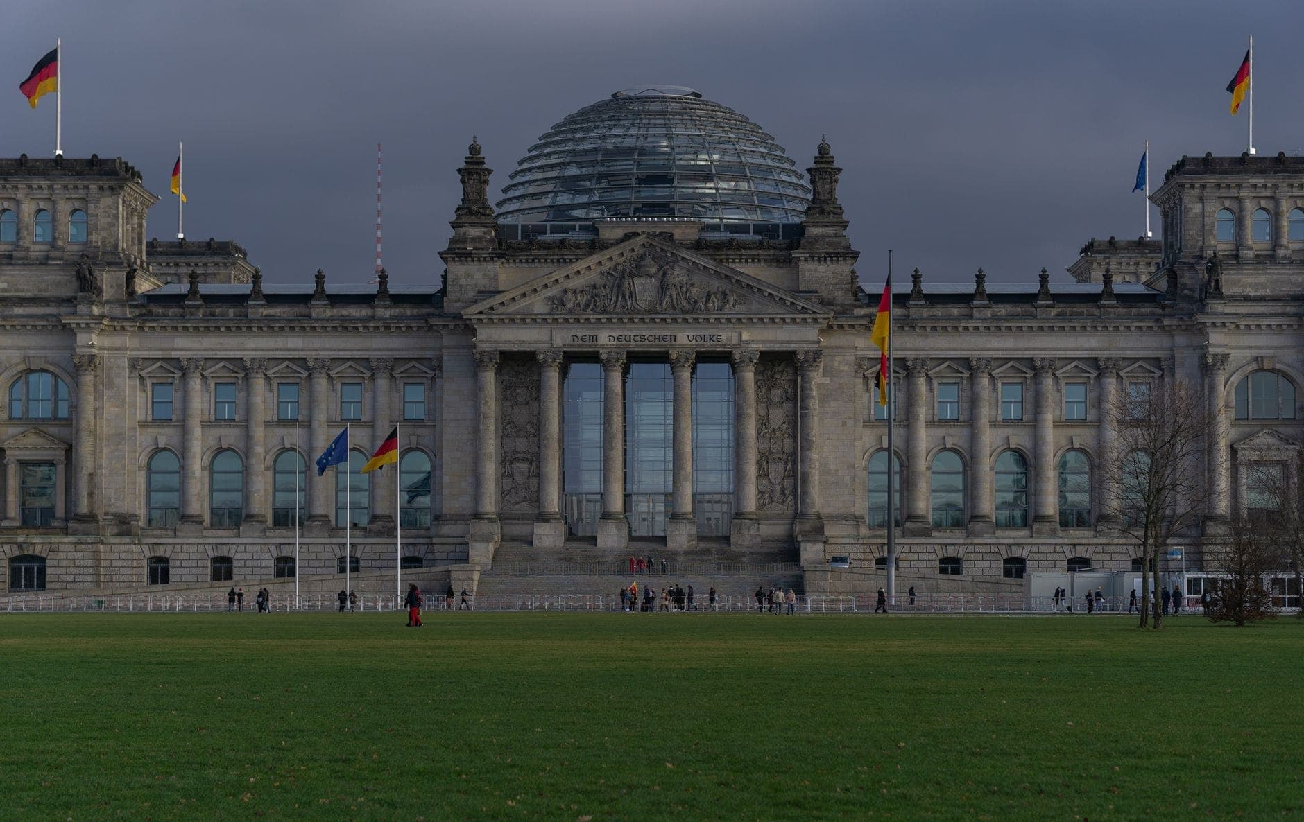 Vista ampia dell'edificio del Reichstag a Berlino con la sua cupola di vetro, bandiere tedesche e persone davanti sotto un cielo nuvoloso.