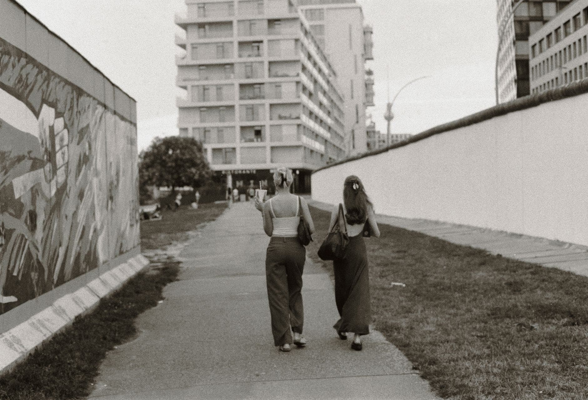 Zwei Personen, die entlang der East Side Gallery in Berlin spazieren, mit Wandmalereien und Stadtgebäuden im Hintergrund.