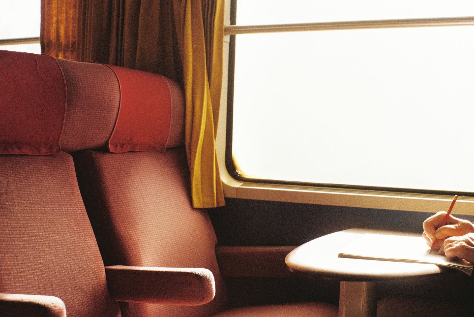 Interno di un confortevole vagone ferroviario a lunga percorrenza con sedili rossi, un tavolo e una persona che scrive vicino al finestrino.