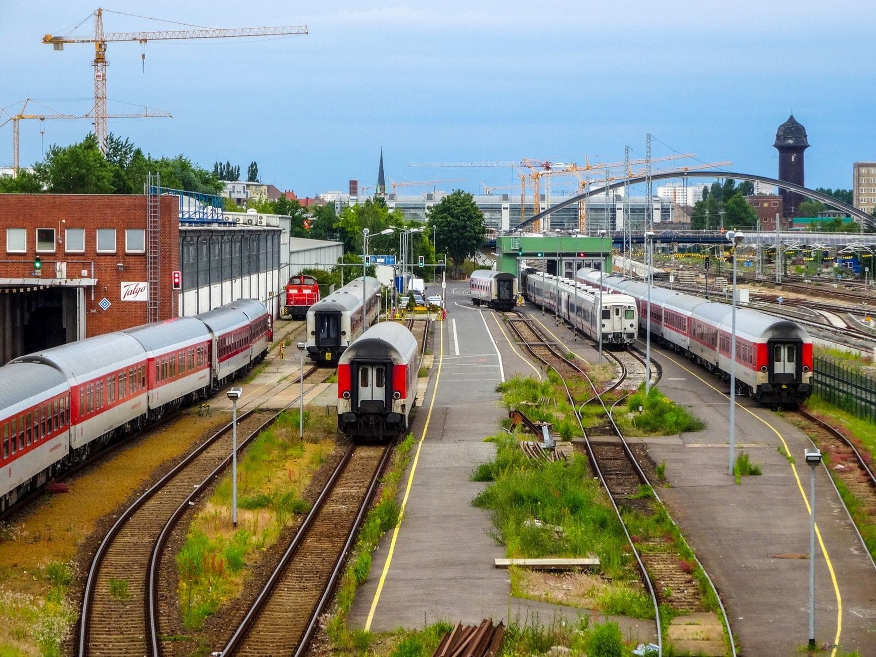 ขบวนรถไฟ EuroCity หลายขบวนในลานรางที่เบอร์ลิน มีรางรถไฟ ชานชาลา และเส้นขอบฟ้าเมืองภายใต้ท้องฟ้าสีฟ้า