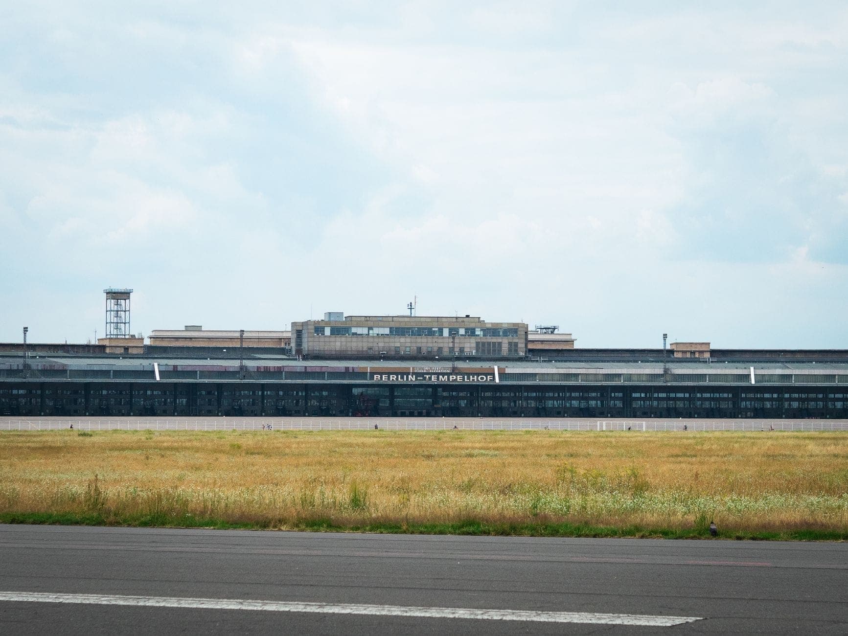 Vista ampia dell'edificio dell'aeroporto Berlin Tempelhof con il suo cartello visibile attraverso un campo aperto erboso e il piazzale.