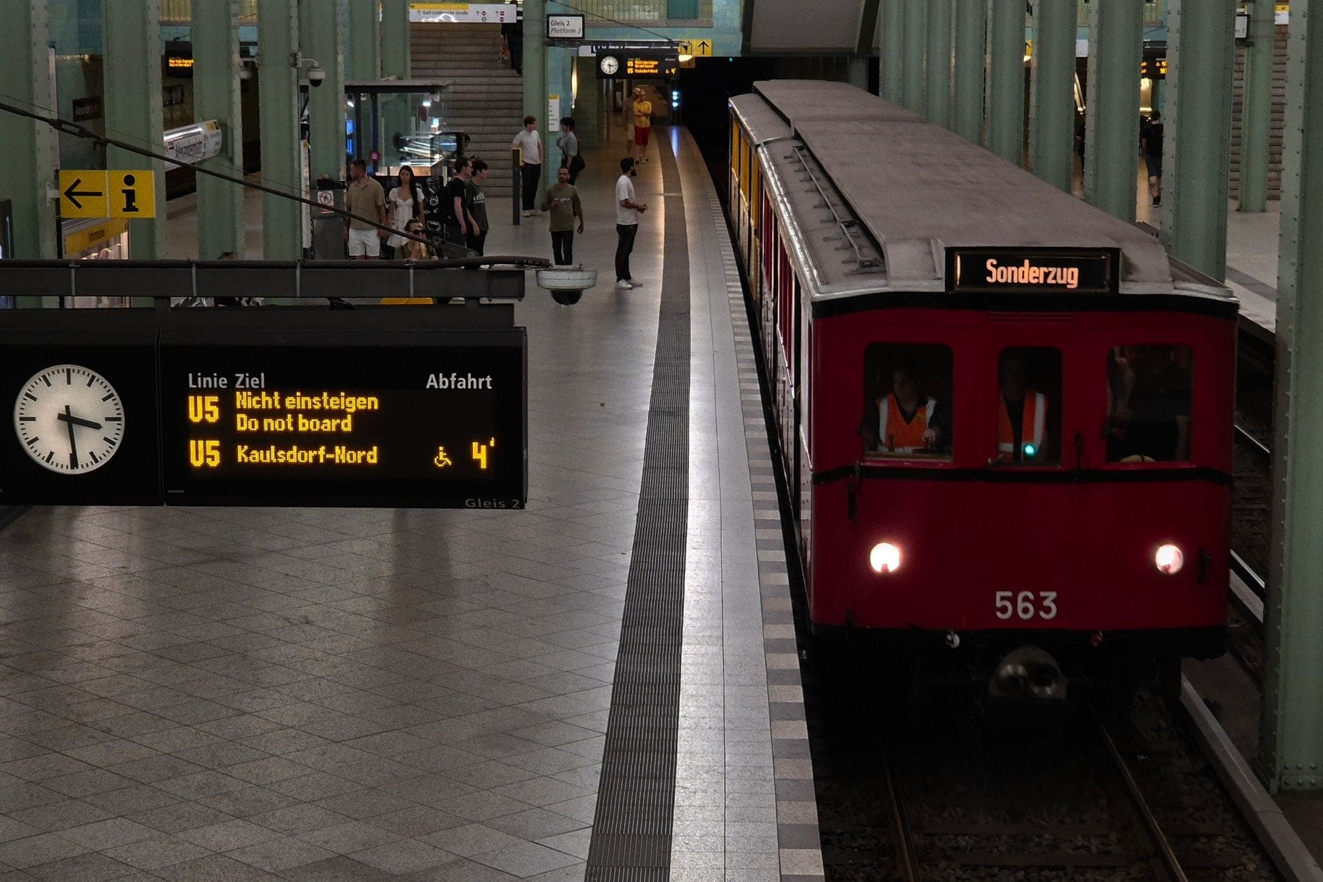สถานีรถไฟใต้ดินในเบอร์ลินพร้อมรถไฟ U-Bahn สีแดง ตารางเวลาแบบดิจิทัล นาฬิกา และผู้โดยสารที่รอบนชานชาลา