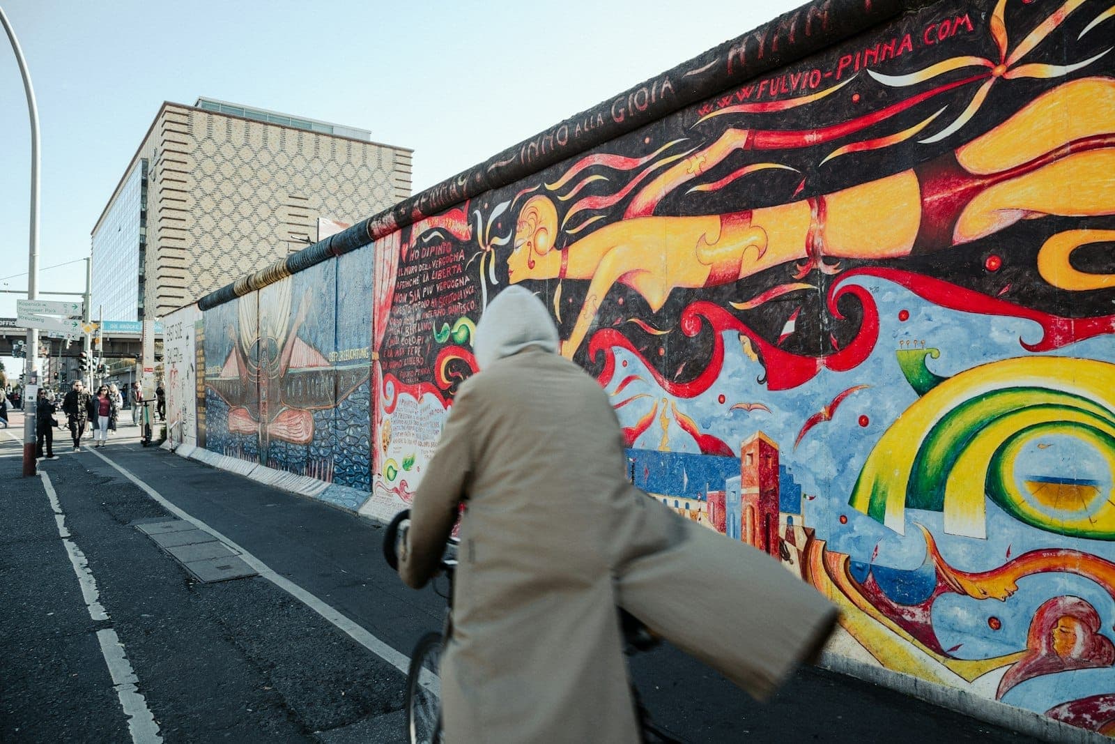 นักปั่นจักรยานสวมเสื้อโค้ทสีเบจขี่ผ่าน Berlin Wall Memorial ที่ระบายสีสดใส โดยมีภาพจิตรกรรมฝาผนังหลากสีสันและอาคารสมัยใหม่เป็นฉากหลังใต้ท้องฟ้าแจ่มใส