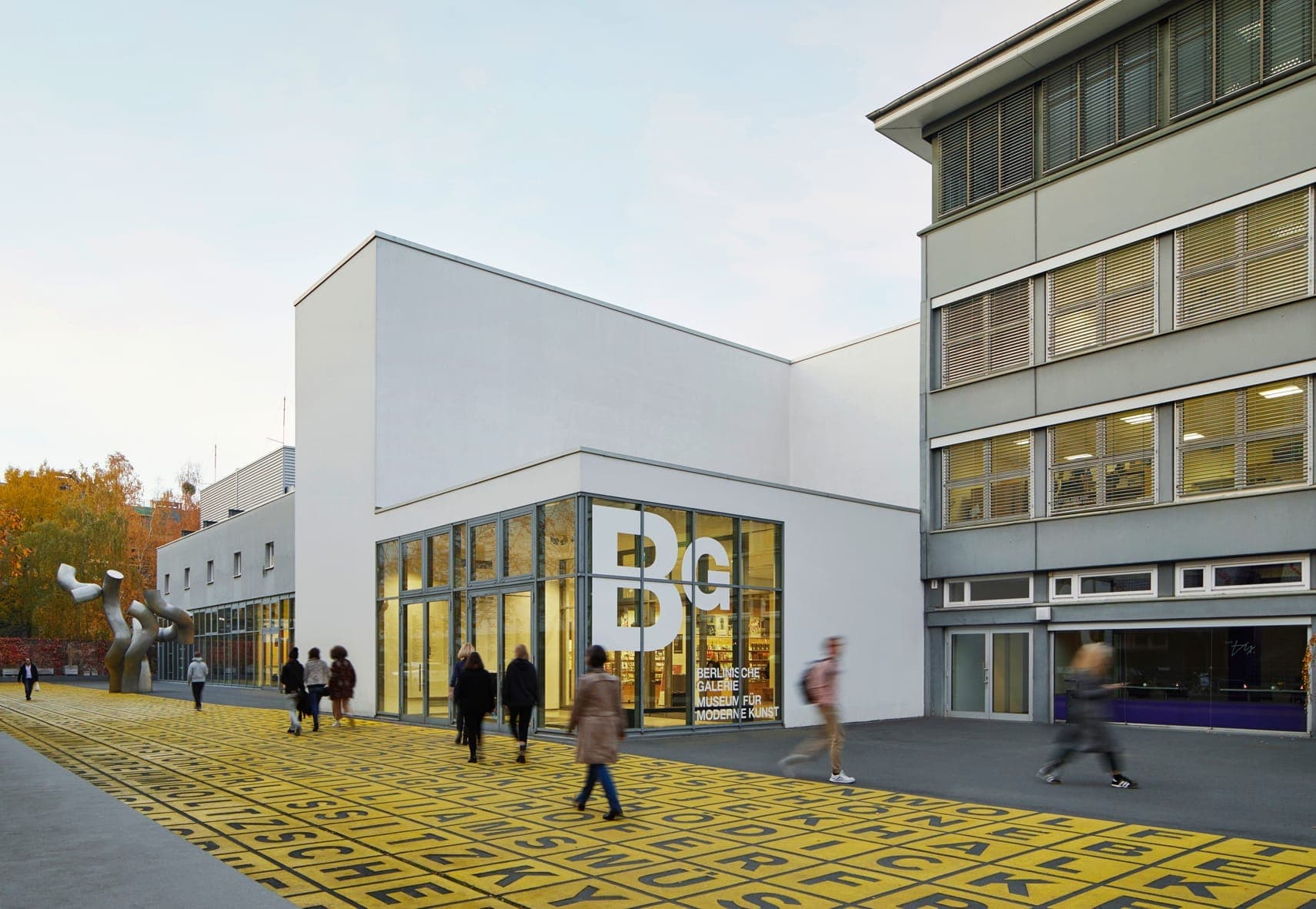ด้านนอกของพิพิธภัณฑ์ Berlinische Galerie ที่มีด้านหน้าอาคารสีขาวทันสมัย หน้าต่างบานใหญ่ และผู้คนเดินบนทางเดินลายสีเหลือง
