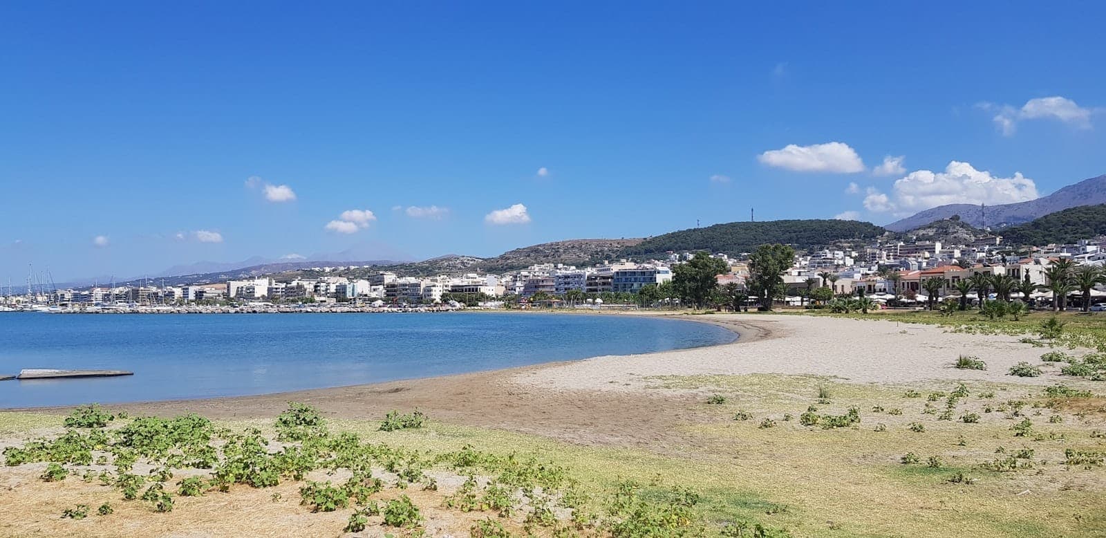 Ampia spiaggia sabbiosa con acqua cristallina, edifici della città e colline sullo sfondo sotto un cielo blu limpido a Rethymno, Creta.