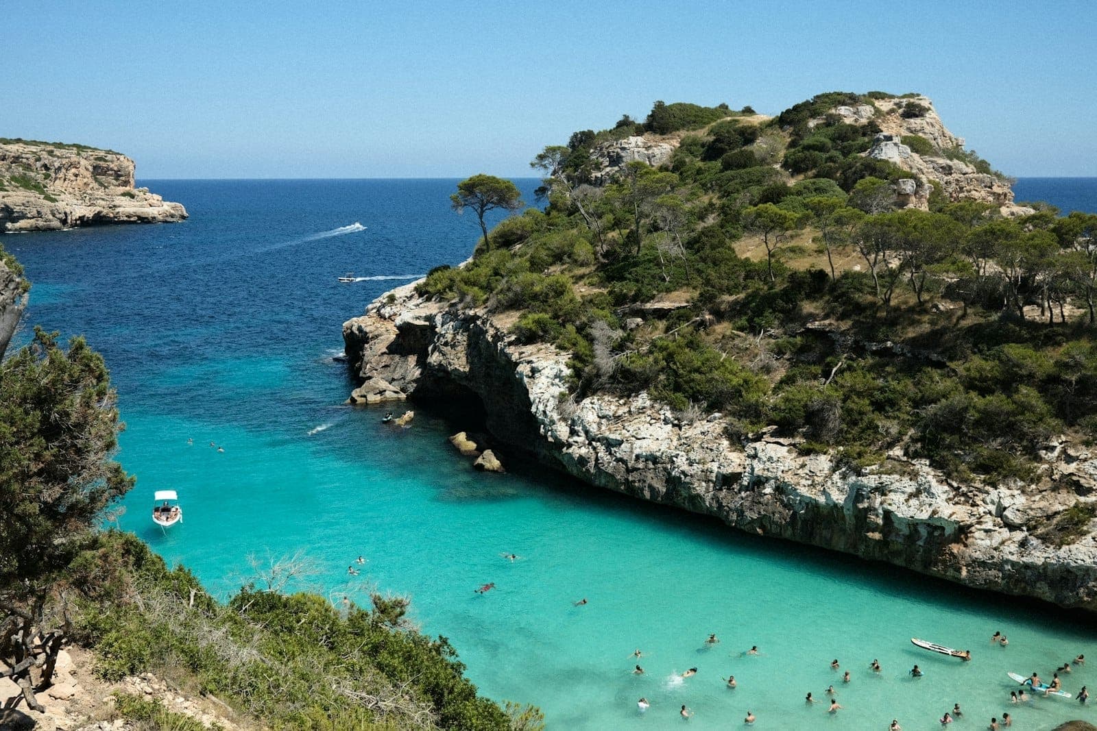 Dramatische Felsbucht auf Mallorca mit türkisblauem Wasser, Schwimmern, Booten und sattgrünen Hügeln unter strahlend blauem Himmel. Einladende Strandkulisse.