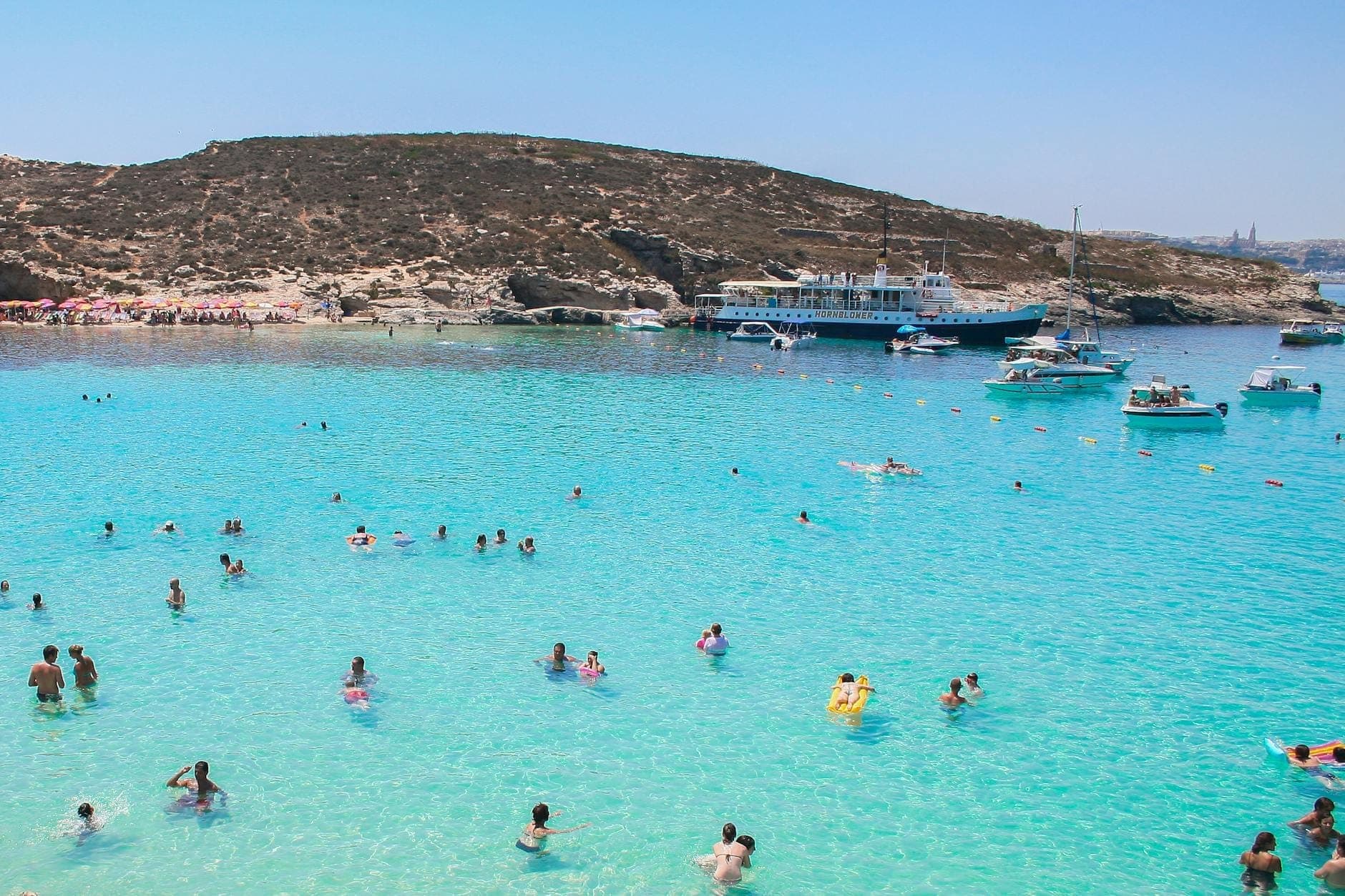 Menschenmassen schwimmen im klaren türkisfarbenen Wasser der Blue Lagoon, Comino, mit Booten und felsigen Hügeln im Hintergrund.