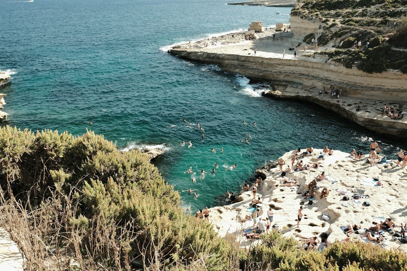 Menschen, die sich auf flachen Kalksteinfelsen sonnen und in natürlichen Felspools an einer maltesischen Küste schwimmen.