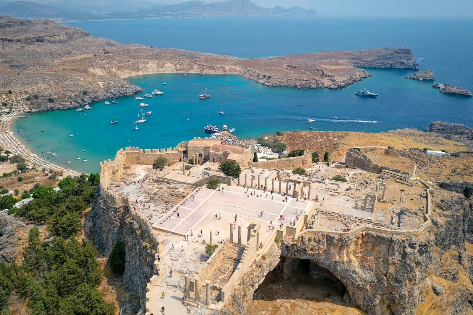 Vue aérienne de l'Acropole de Lindos perchée sur une falaise au-dessus d'une baie turquoise, avec un littoral spectaculaire et des bateaux amarrés à Rhodes, Grèce.