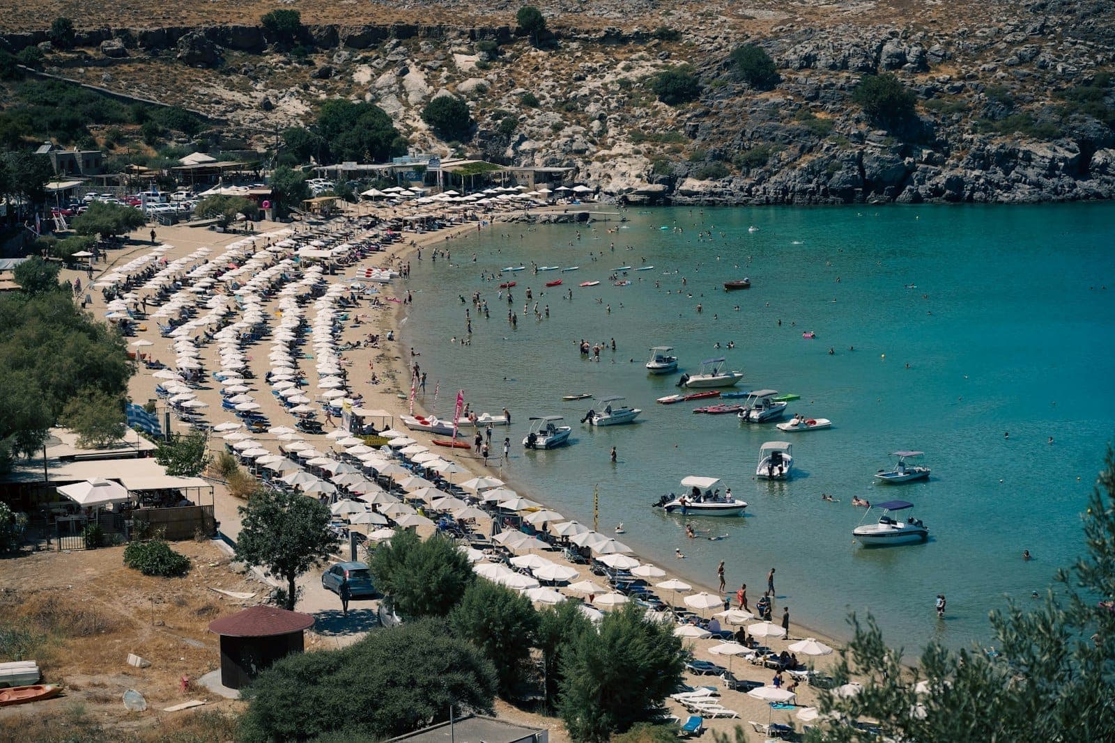 Vue aérienne d'une plage de sable à Rhodes bordée de parasols, d'eaux turquoise peu profondes et de familles profitant de la mer avec des bateaux à proximité.