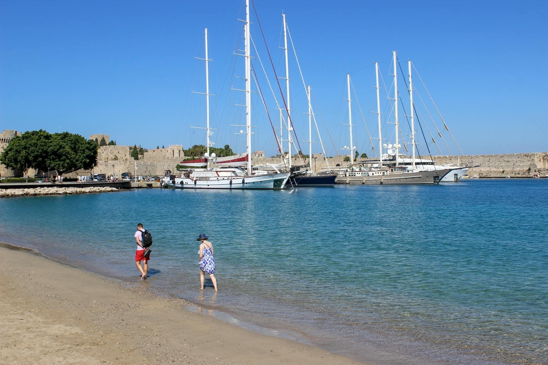 Petite plage de sable avec deux personnes se promenant, eau bleue cristalline, yachts et fortifications médiévales en arrière-plan à Rhodes.