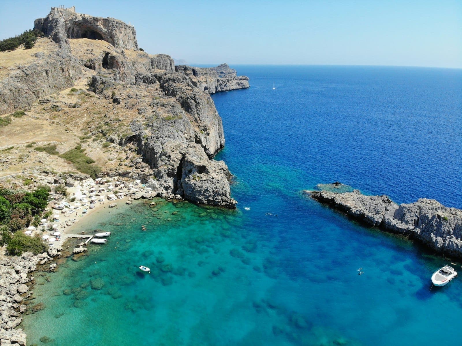 Une large crique de sable aux eaux turquoise, dominée par des falaises spectaculaires et l'Acropole de Lindos, avec bateaux et baigneurs sur le célèbre littoral de Rhodes.