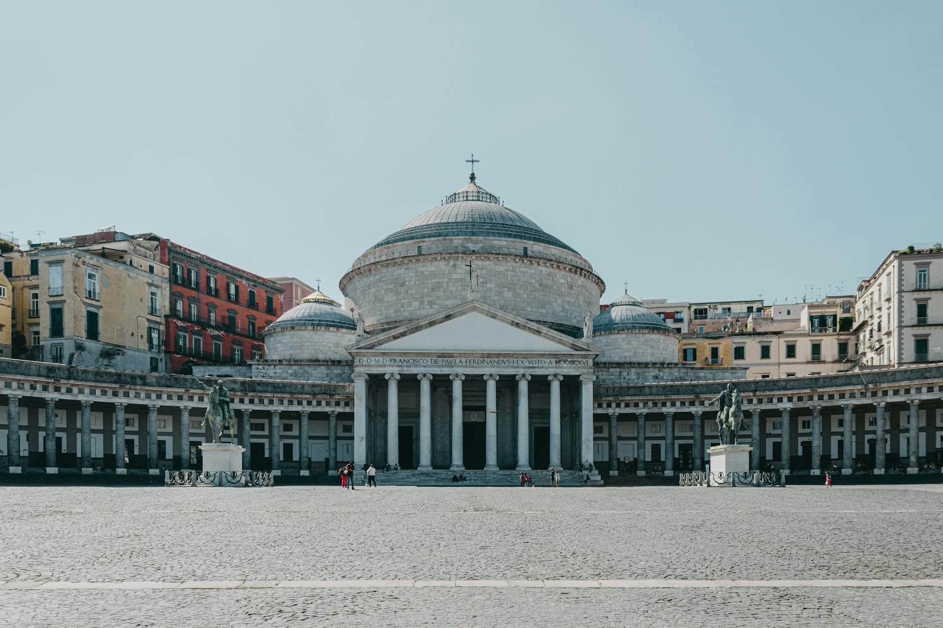Vista ampla da grande Basílica de San Francesco di Paola com cúpula na Piazza del Plebiscito de Nápoles, com alas com colunatas e edifícios coloridos.