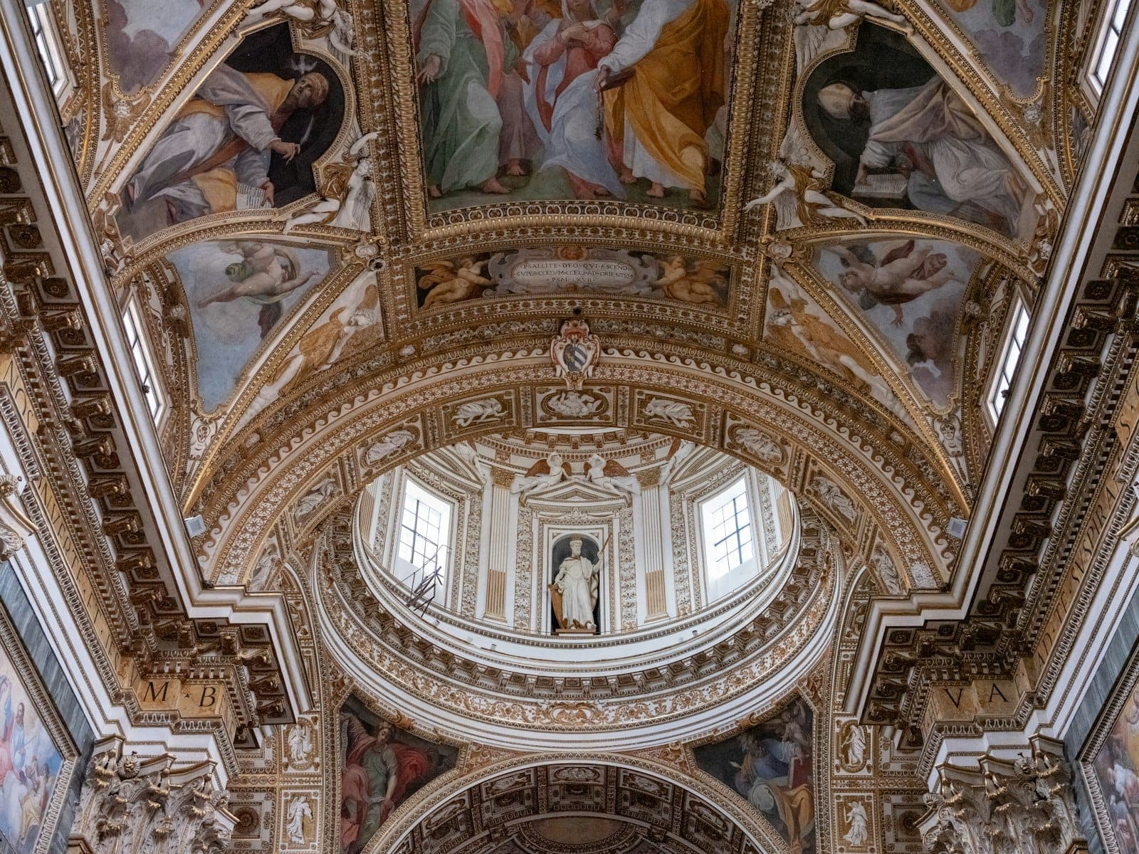 Interior de igreja barroca ricamente decorado em Roma com arcos dourados, estuques ornamentados e coloridos afrescos no teto vistos de baixo.