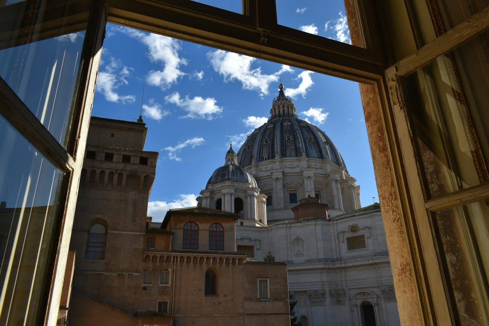 Vista da cúpula da Basílica de São Pedro através de uma janela aberta, com céu azul e nuvens sobre os telhados de Roma.