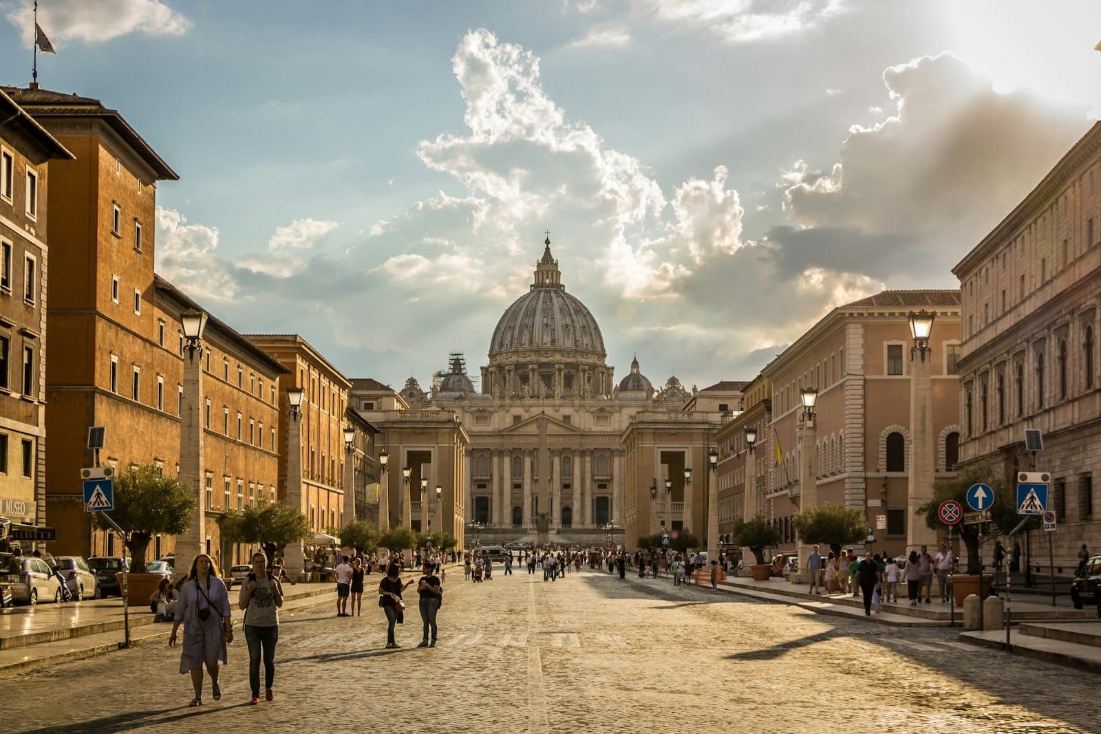 Vista ampla da Basílica de São Pedro com sua icônica cúpula ao final de uma movimentada rua em Roma, com pessoas caminhando em direção ao ponto turístico.