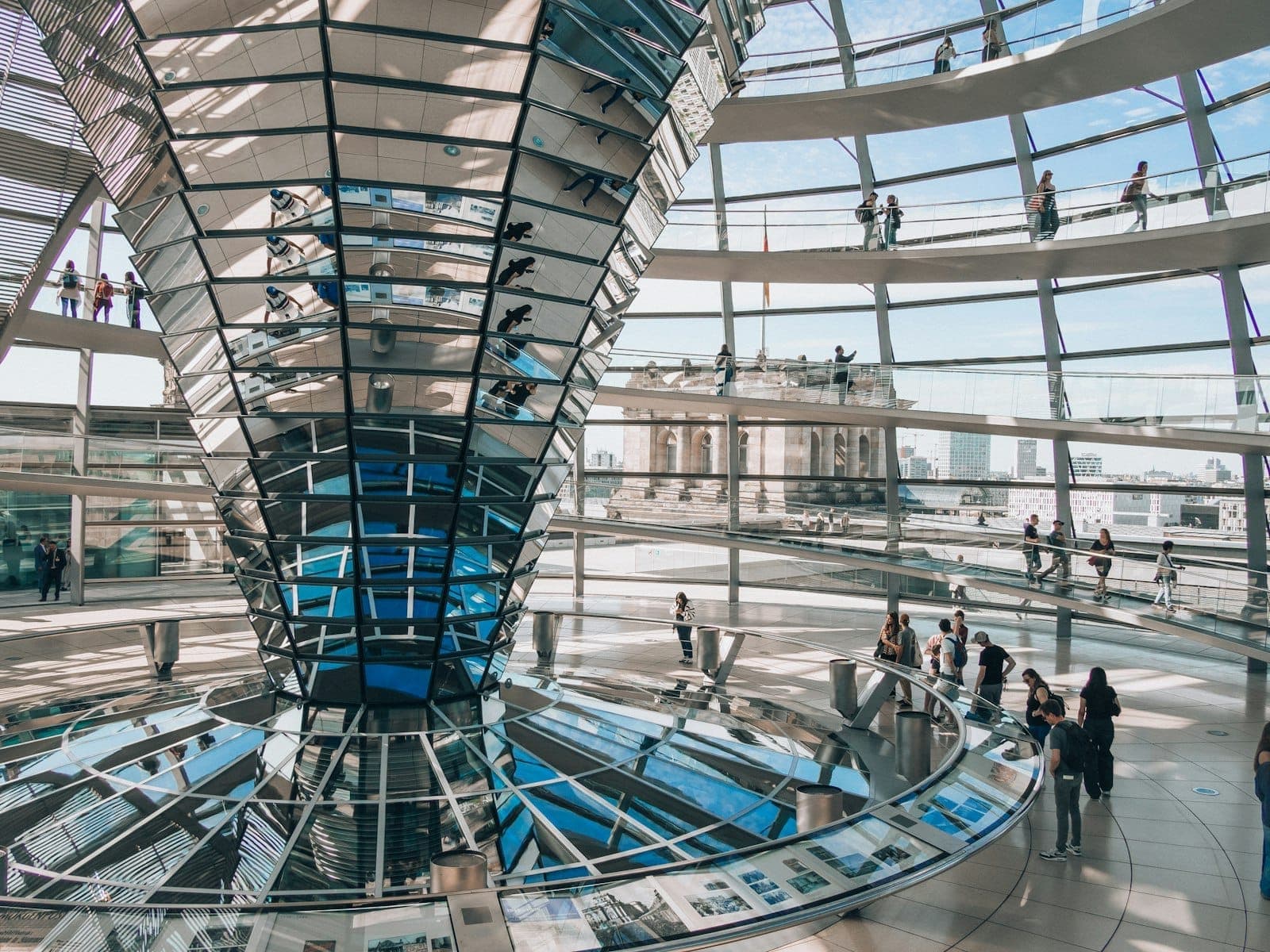 Szeroki widok na szklanie sklepienie wnętrza berlińskiego Reichstagu z odwiedzającymi spacerującymi po zakrzywionych rampach i wpadającym naturalnym światłem.