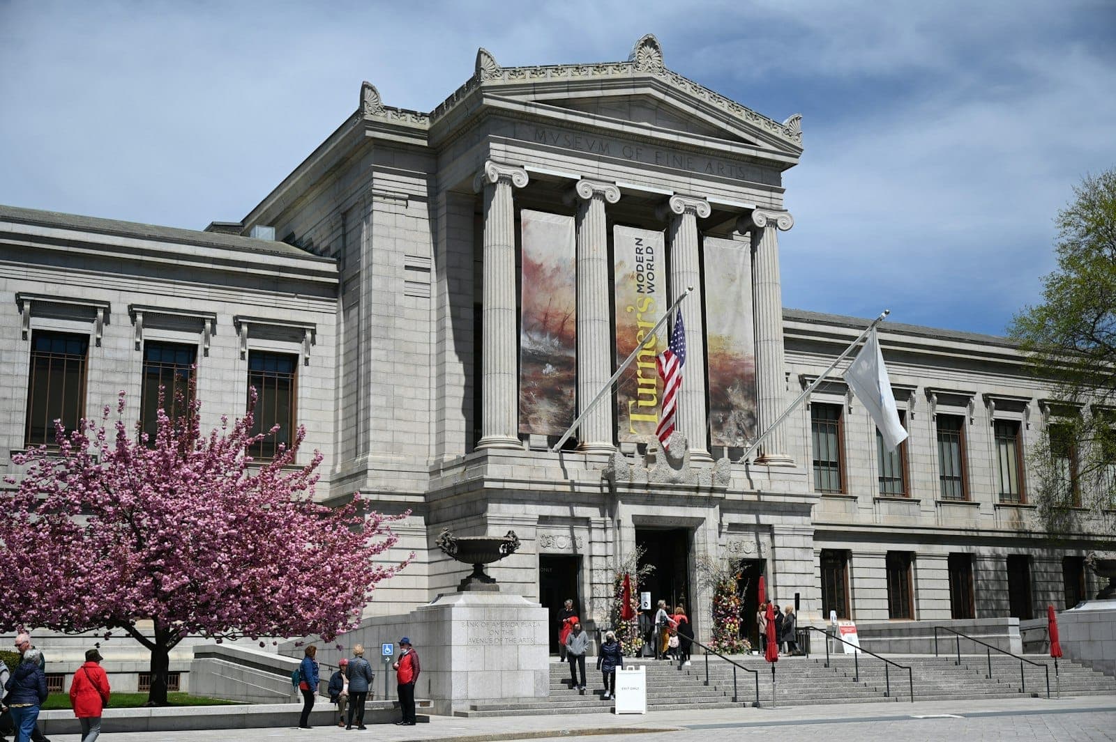Vue de face du Museum of Fine Arts Boston avec colonnes, bannières, arbre en fleurs et personnes à l'extérieur par une journée ensoleillée.