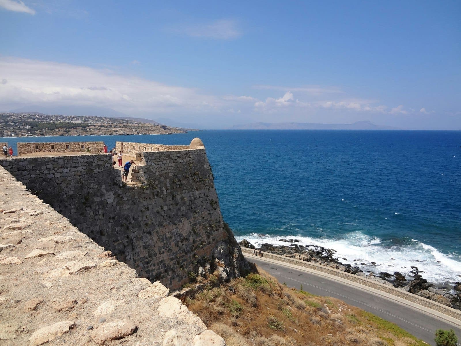 Muro di fortezza marittima in pietra su una scogliera che si affaccia sul blu del Mediterraneo, con visitatori e una spettacolare costa a Creta.