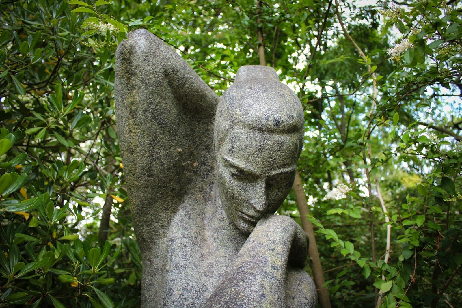Close-up de uma escultura de pedra de uma figura humana, rodeada por folhagem verde em um jardim ao ar livre.